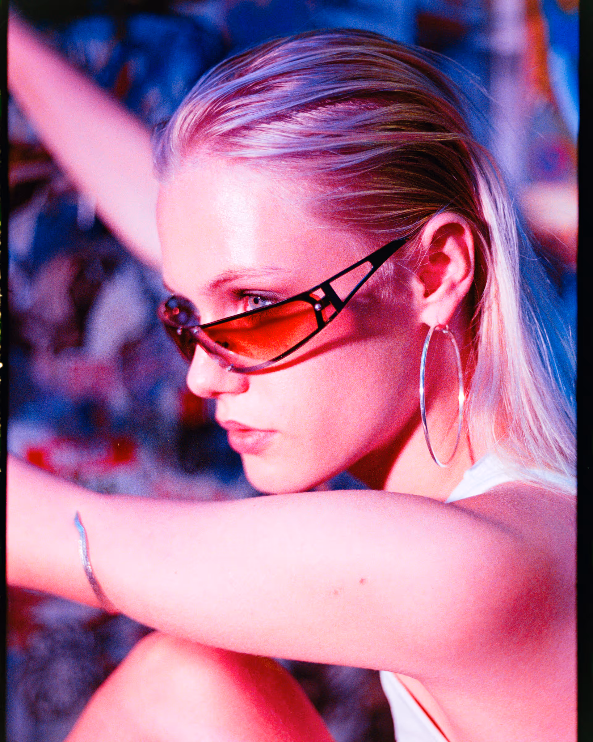 Close-up side profile of a woman with slicked-back blonde hair wearing red-tinted geometric sunglasses and large hoop earrings.