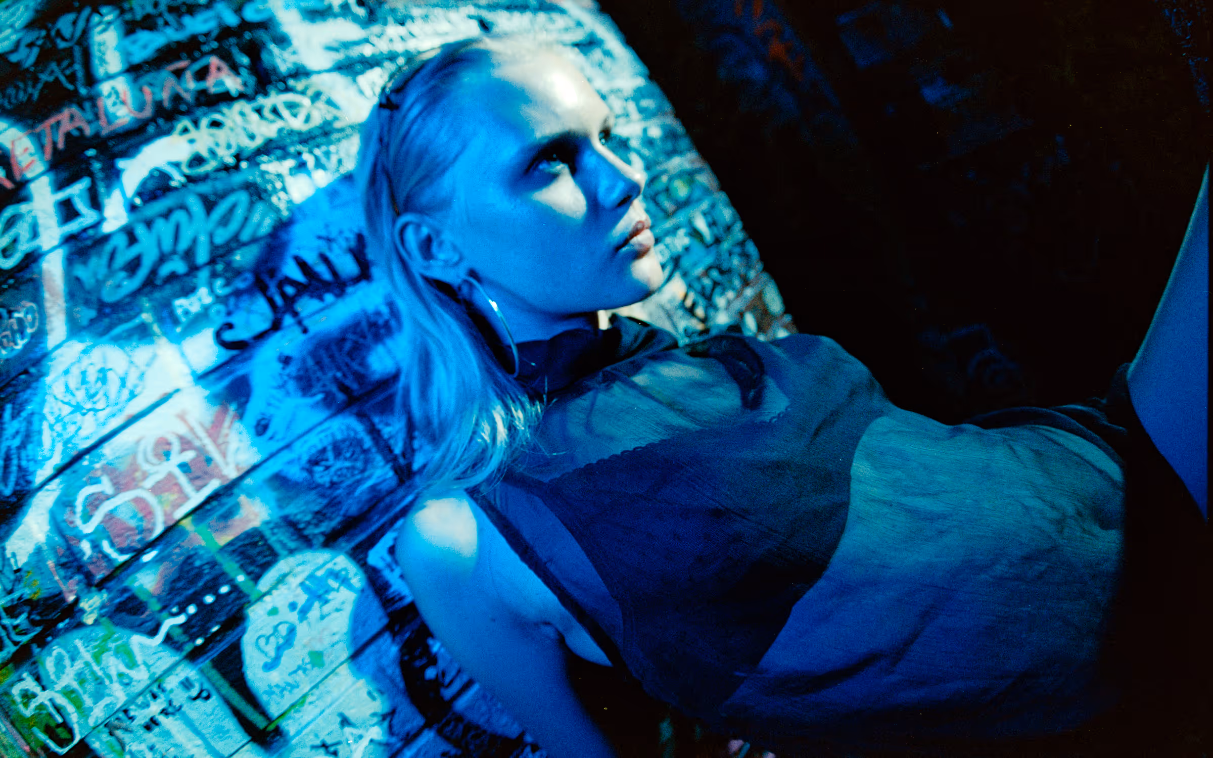 Young woman with blonde hair wearing a sheer black top and large hoop earrings standing against a graffiti-covered wall illuminated by blue light.