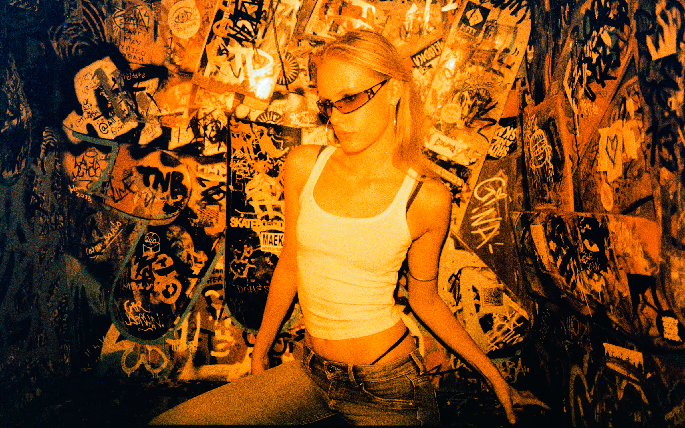 Young woman in sunglasses, white tank top, and jeans posing against a heavily graffiti-covered wall with skateboards.