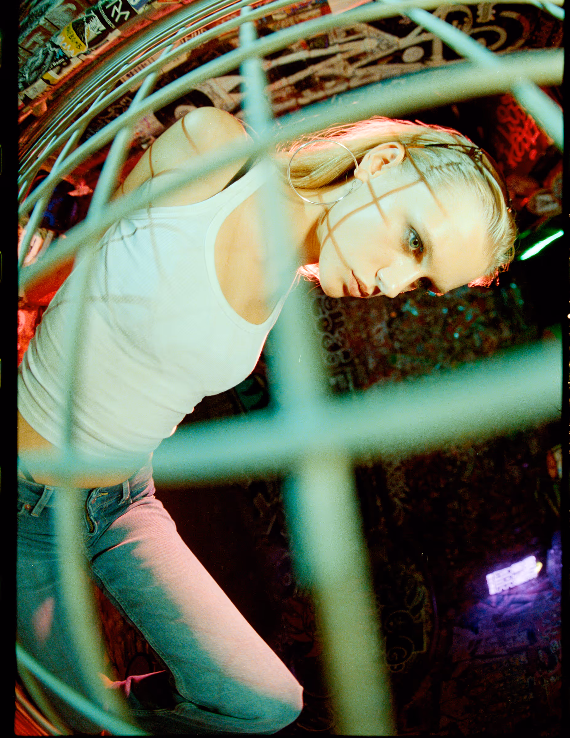 Blonde woman wearing a white tank top and blue jeans leans forward behind a metal grid in a dimly lit graffiti-covered space.