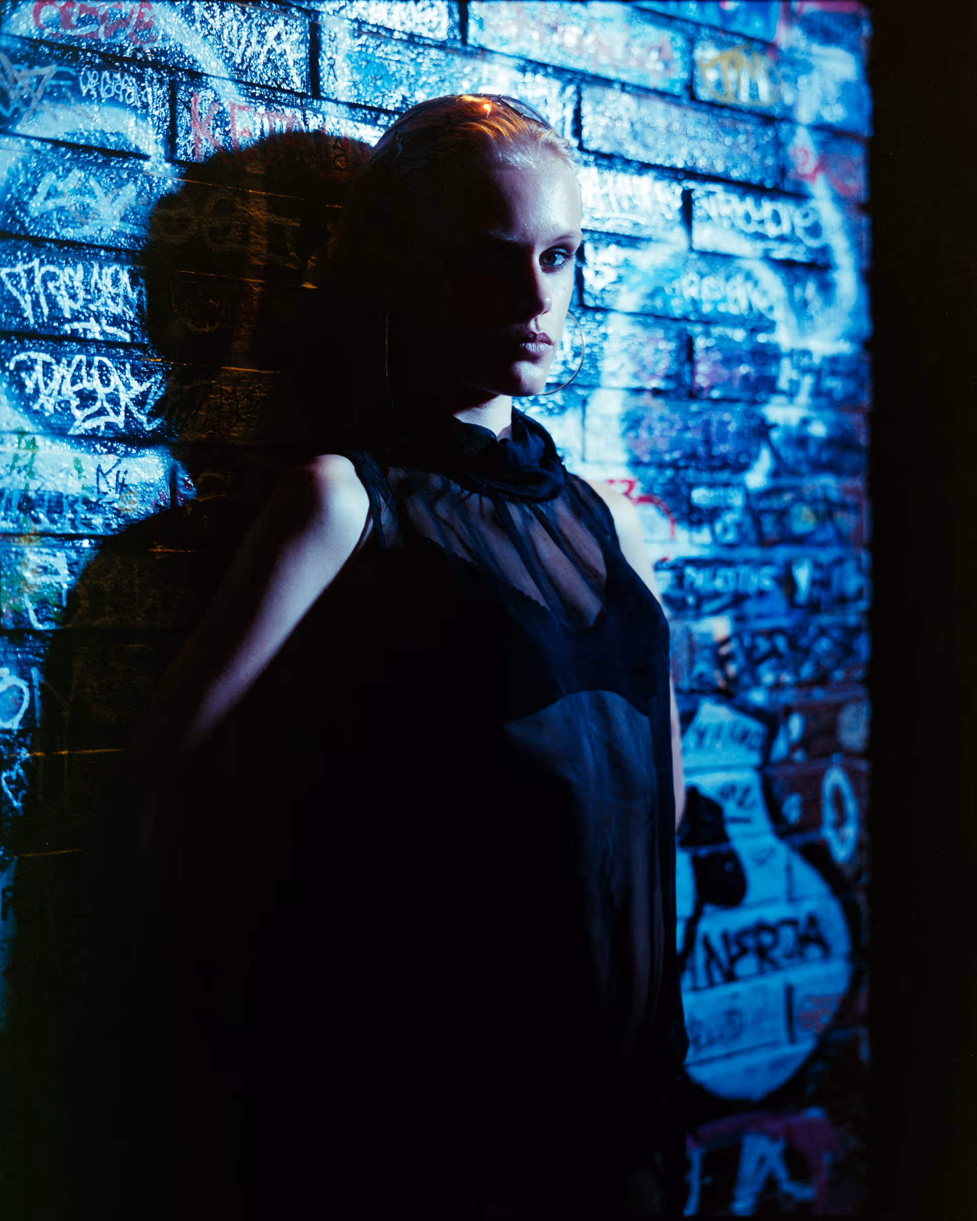 Person with blonde hair and hoop earrings standing against a graffiti-covered brick wall with dramatic blue lighting casting a shadow.