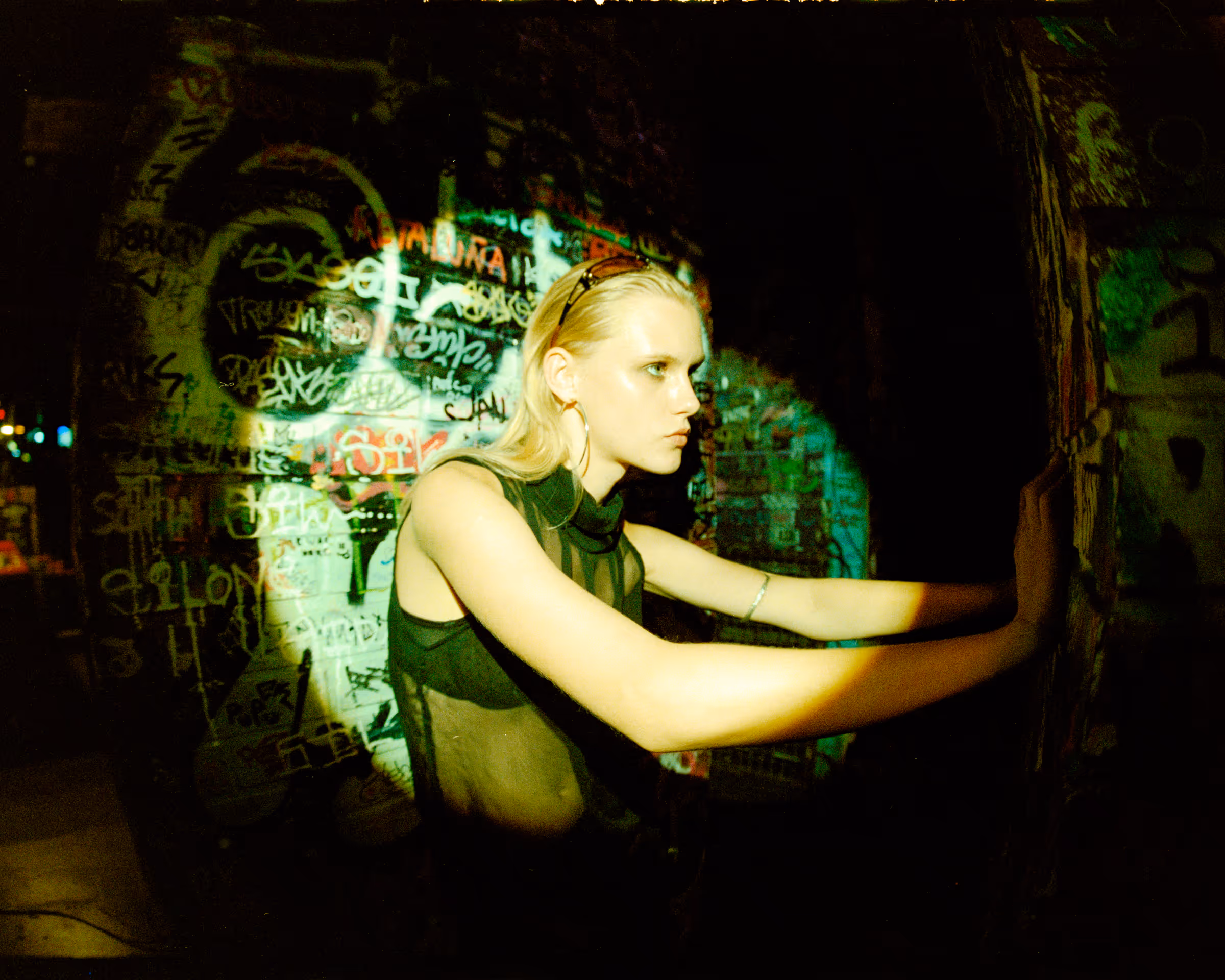Blonde woman in a sleeveless black top leaning with both hands against a graffiti-covered wall, illuminated by a spotlight in a dark urban setting.