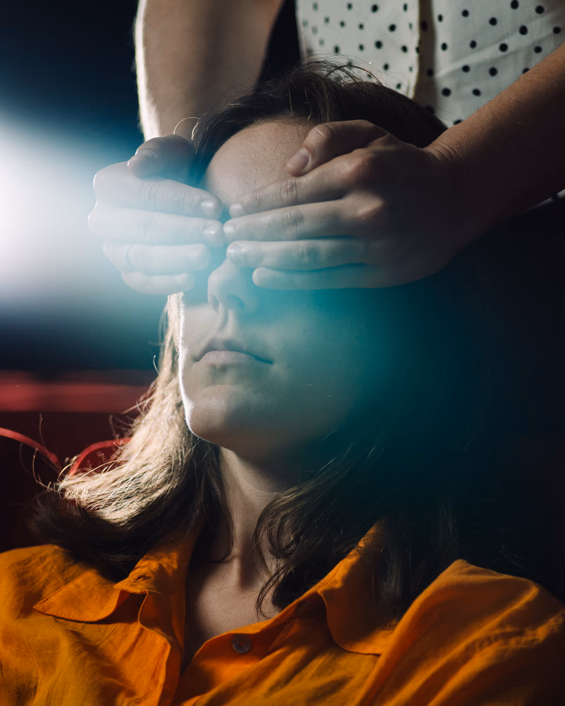 Person with long hair in an orange shirt has their eyes covered by another person's hands.