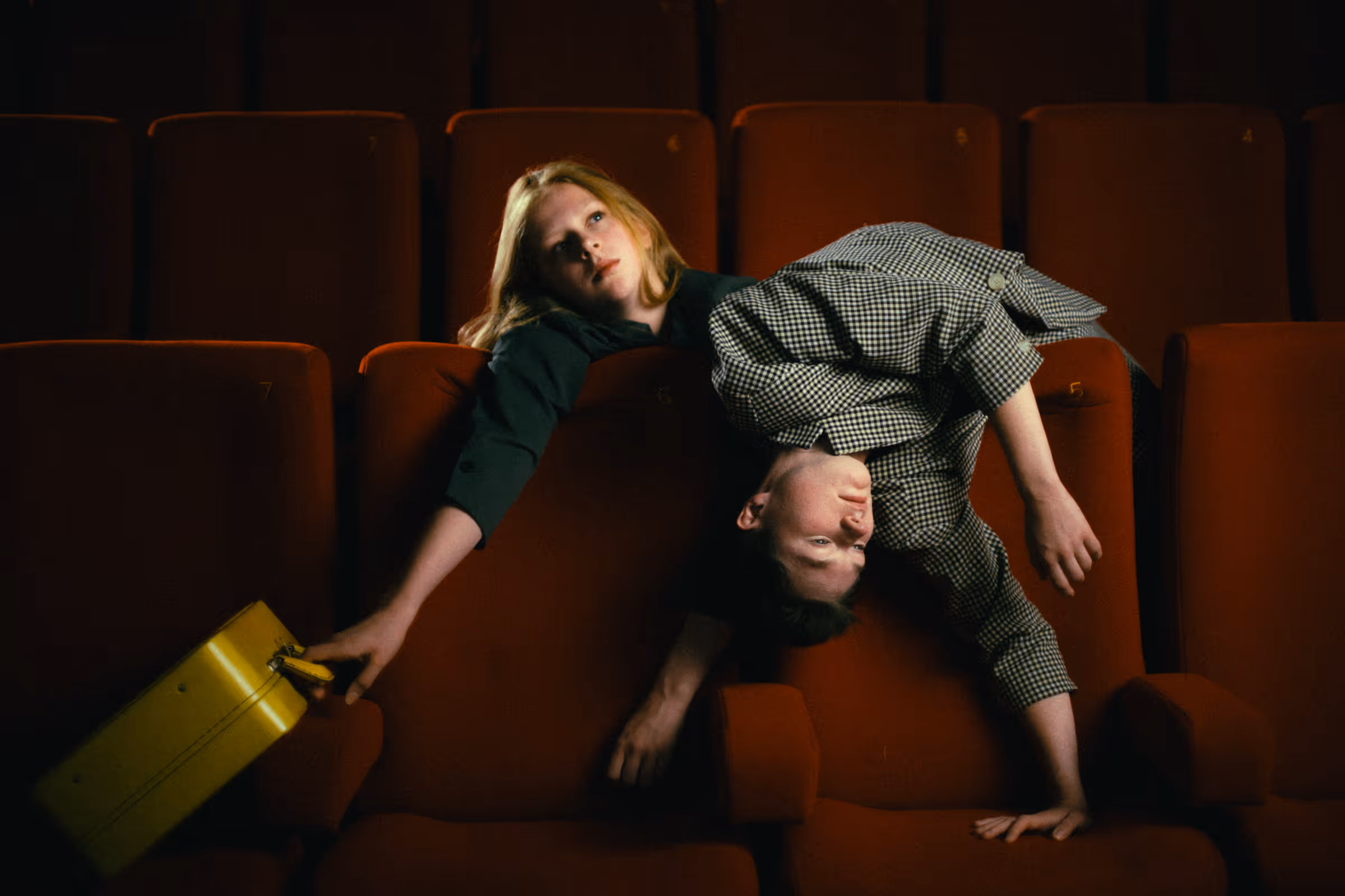 Two people lying across adjacent seats in a dimly lit theater, one holding a yellow briefcase and the other partly draped over them.
