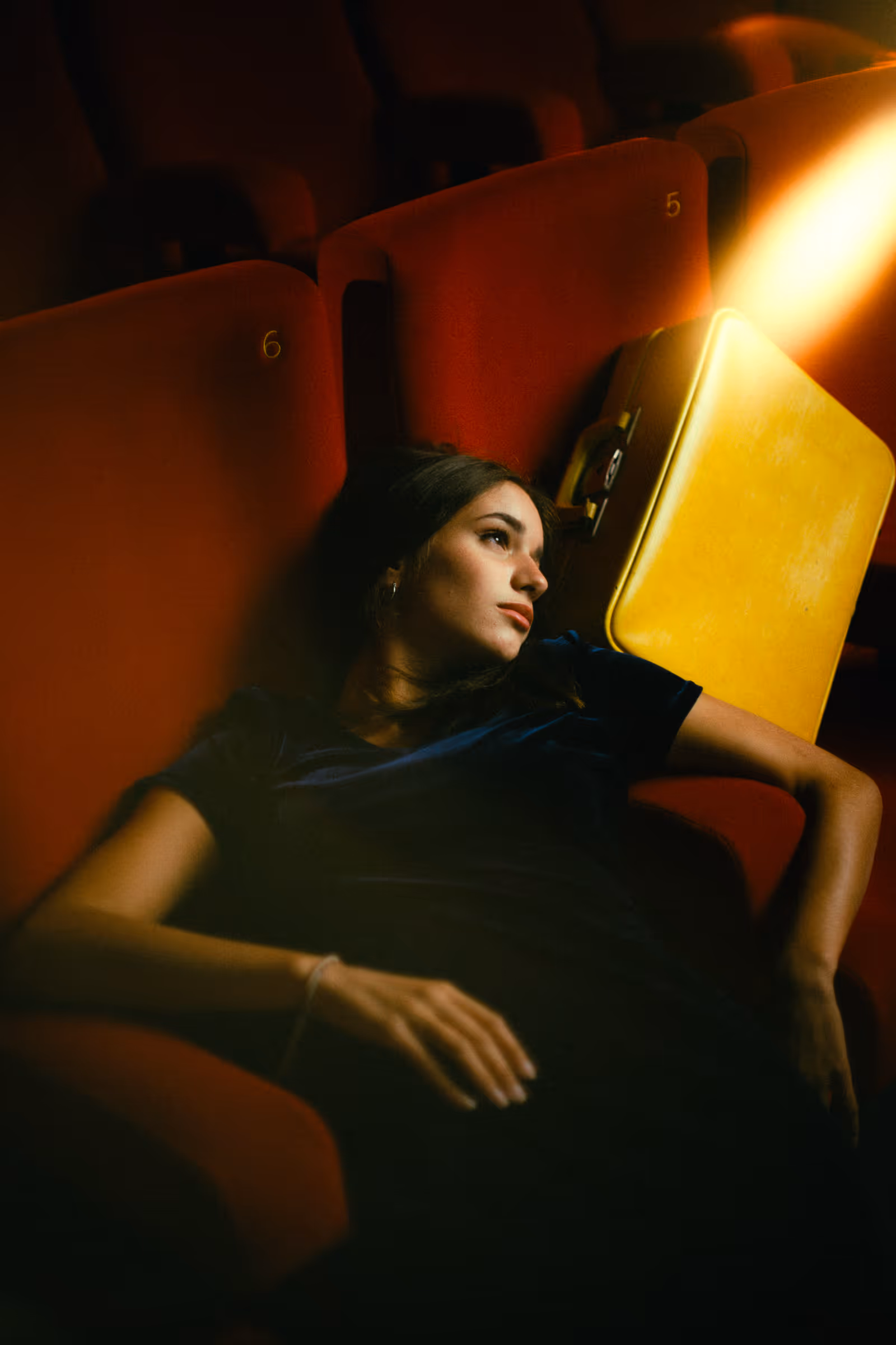 Young woman reclining on red theater seats with a yellow suitcase beside her, looking contemplative.