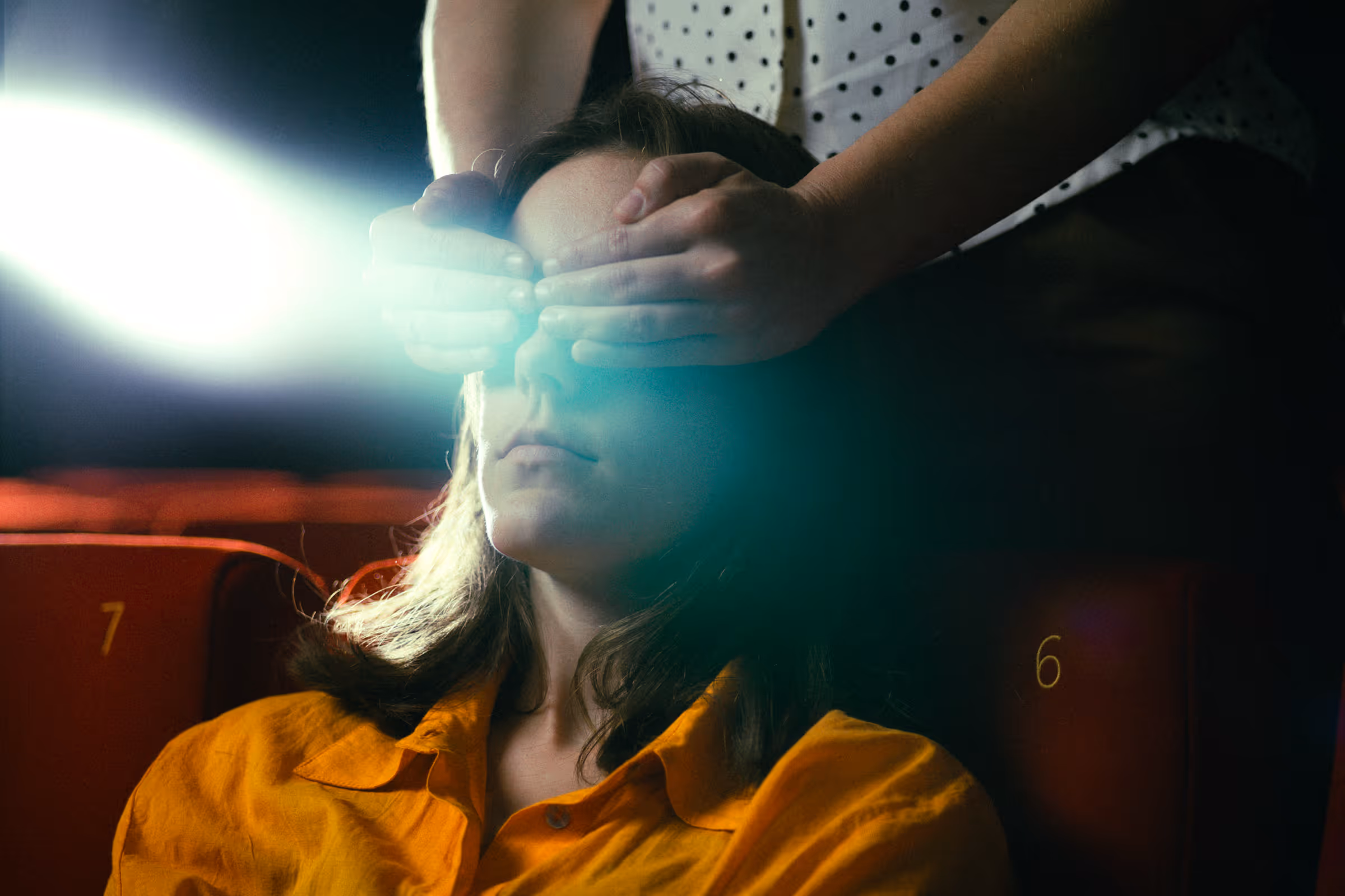 Person in an orange shirt sitting in a theater seat with hands covering their eyes and a bright light shining from the side.