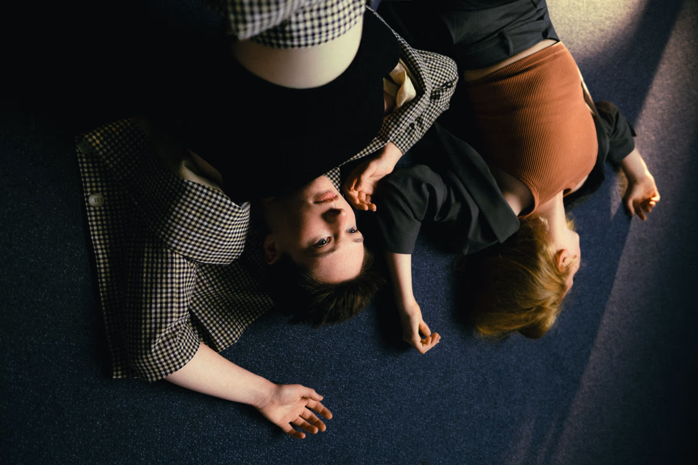 Two people lying on a dark carpeted floor, one with short hair and checkered shirt looking up, the other with long red hair in an orange top facing down.
