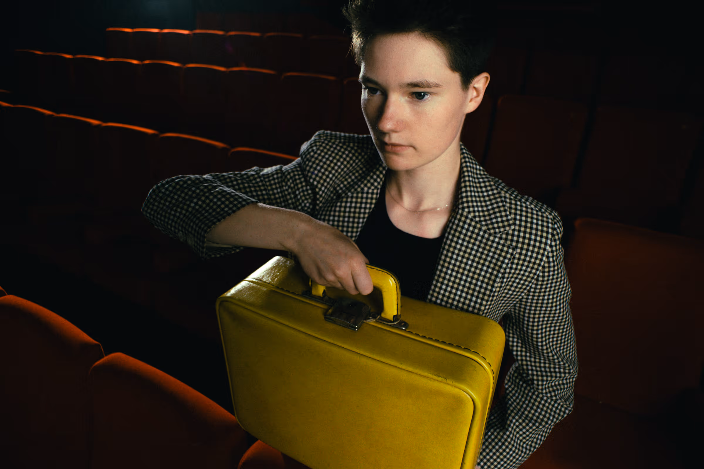 Person with short hair in a checkered blazer holding a bright yellow briefcase in a dark theater with red seats.