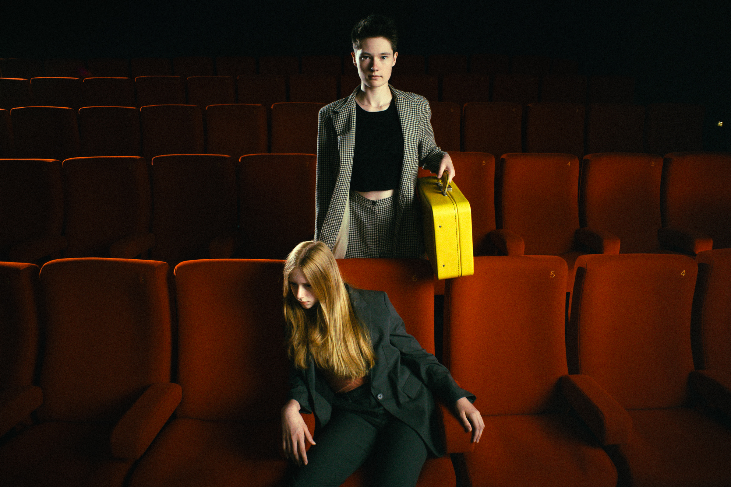 Two young women in a theater with empty red seats, one sitting slouched and the other standing behind with a yellow suitcase.