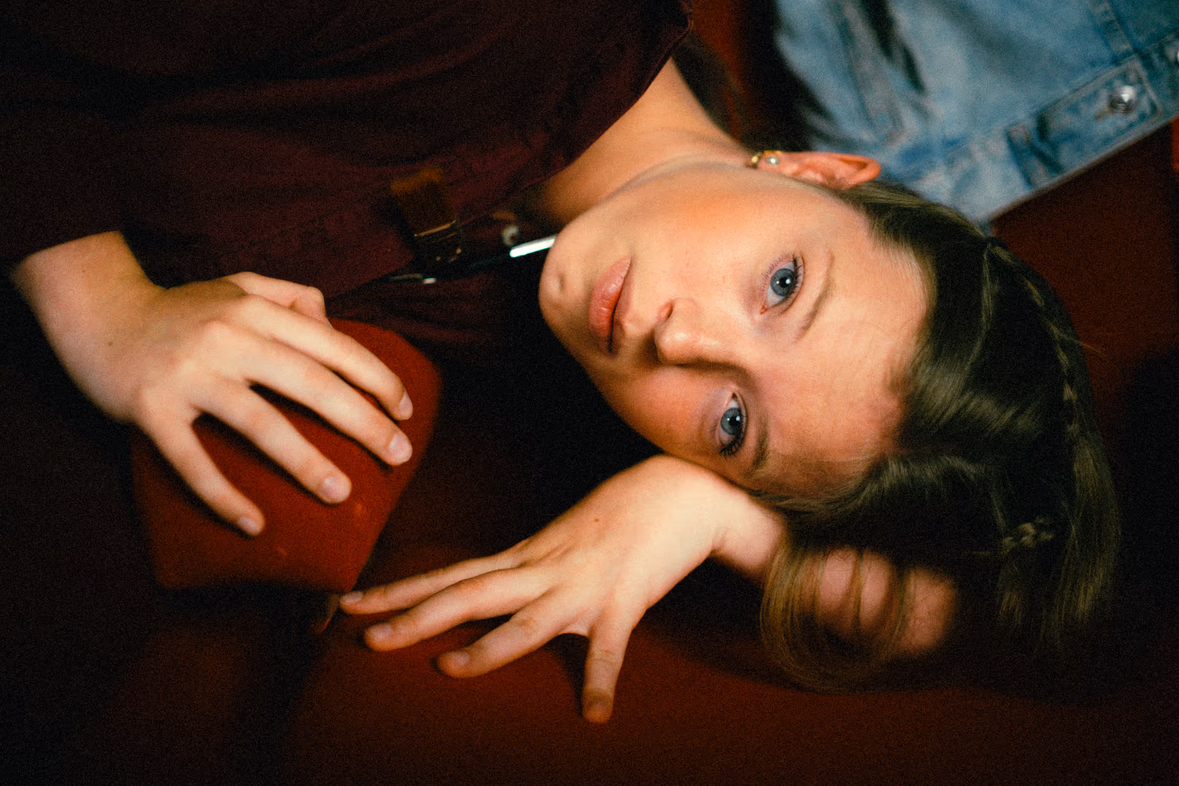 Young woman with blue eyes and braided hair lying on a red surface, resting her head on her arm.