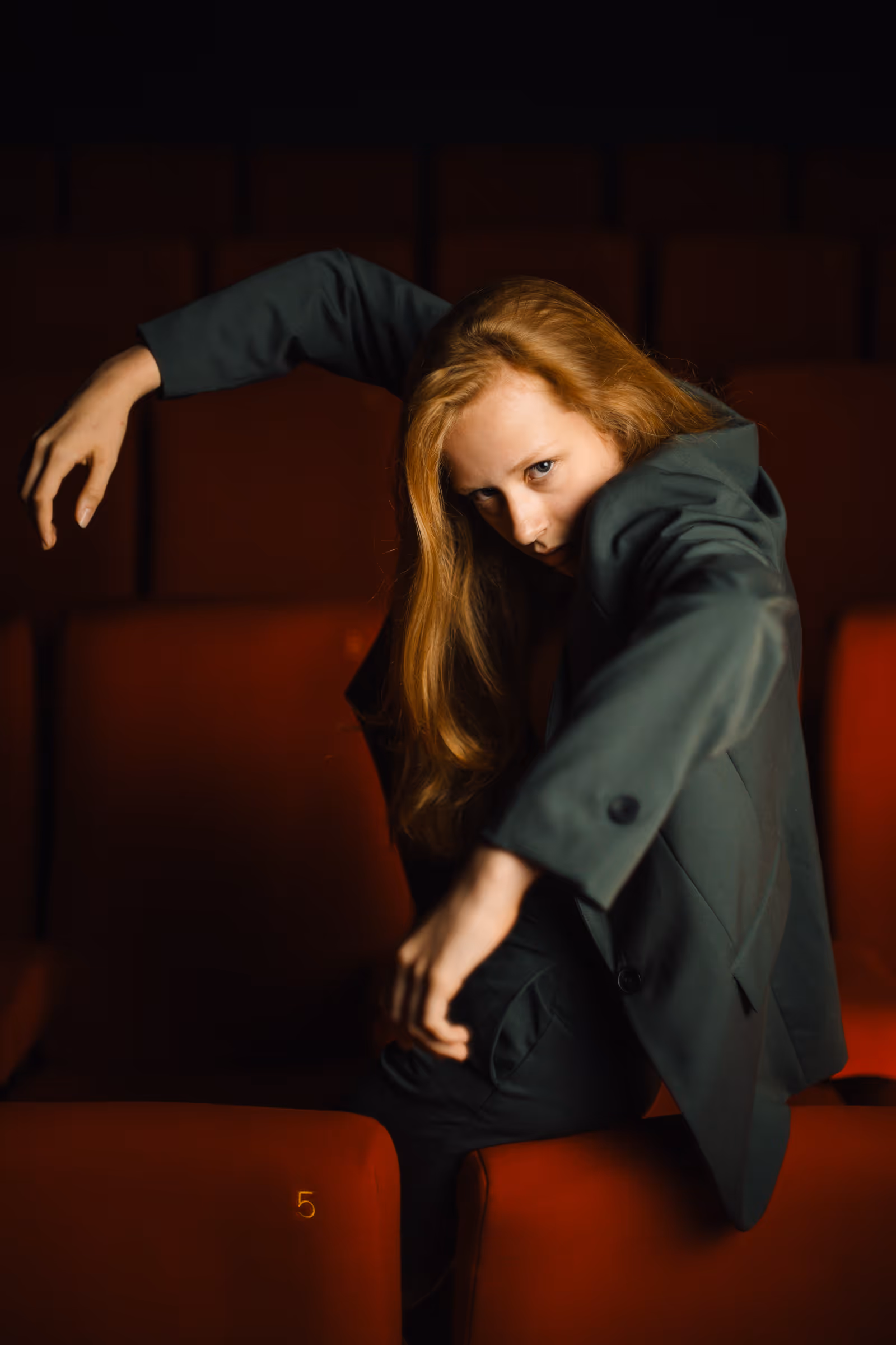 Young woman with long red hair wearing a dark suit jacket striking a dance pose over red theater seats in a dimly lit room.
