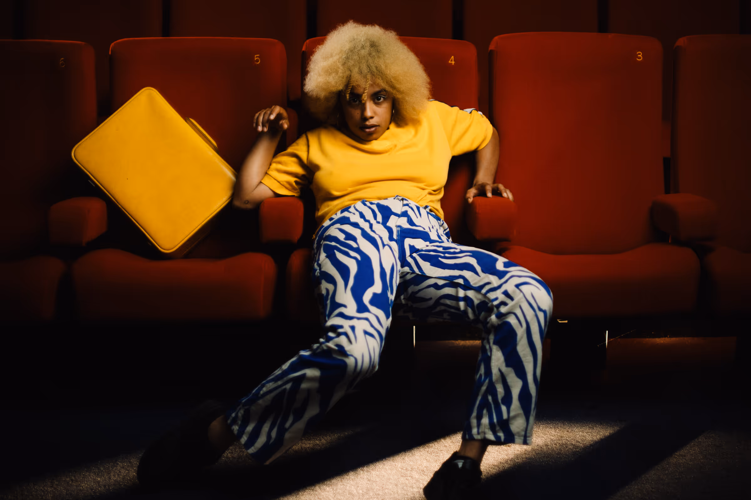 Person with curly blond hair wearing a yellow shirt and blue-white patterned pants lounging across red theater seats with a yellow bag on one seat.