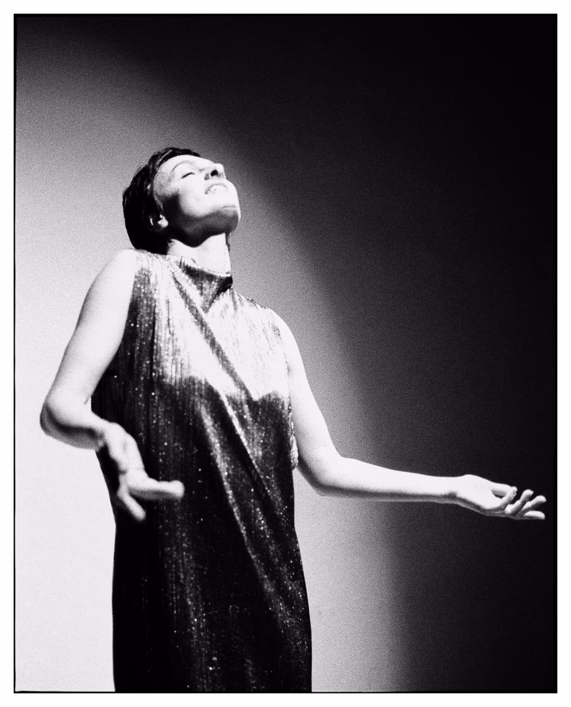 Black and white photo of a woman in a sleeveless shimmering dress with arms outstretched and head tilted back, eyes closed, appearing joyful.