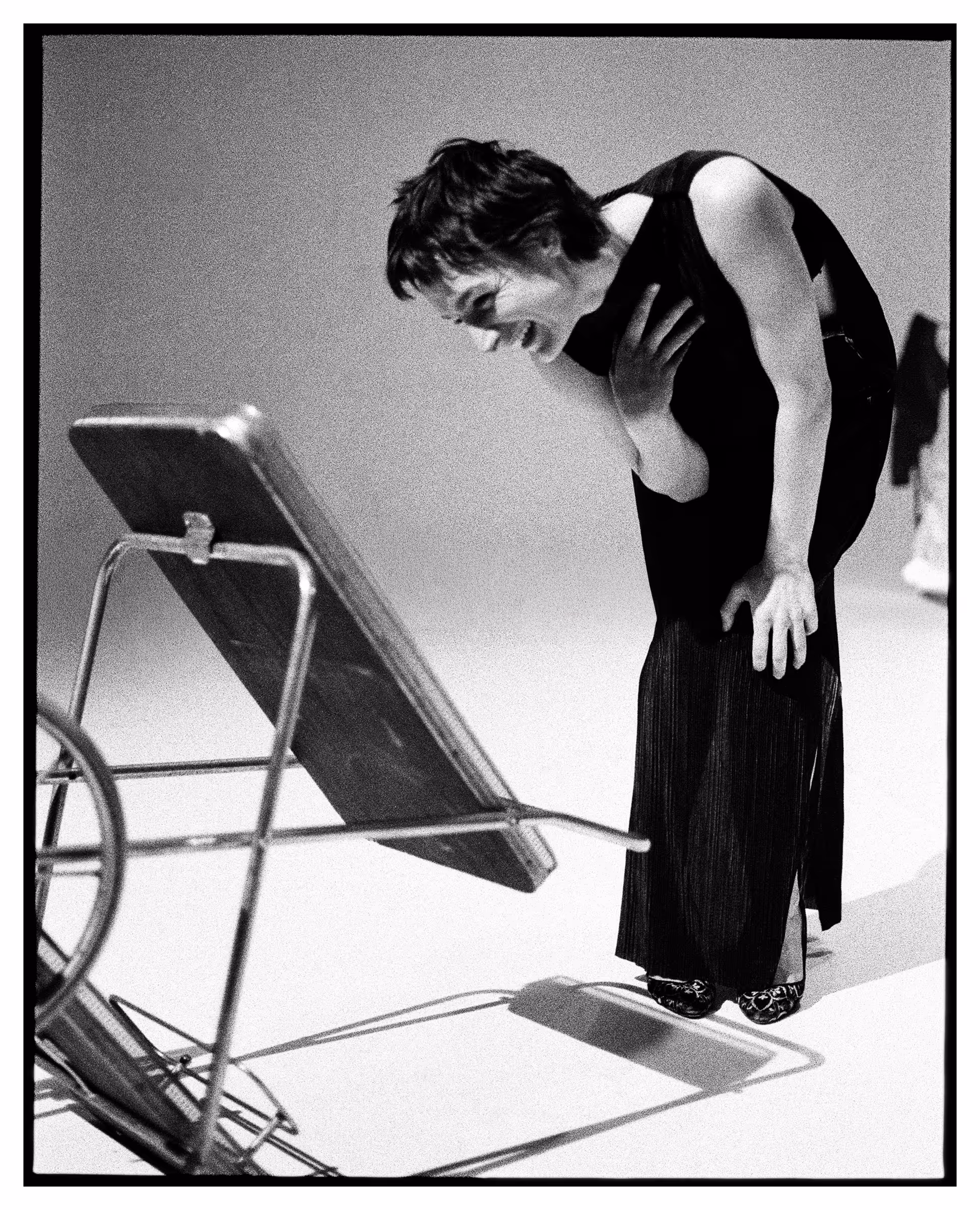Black and white photo of a woman in a long dress bending forward and laughing near an overturned chair.
