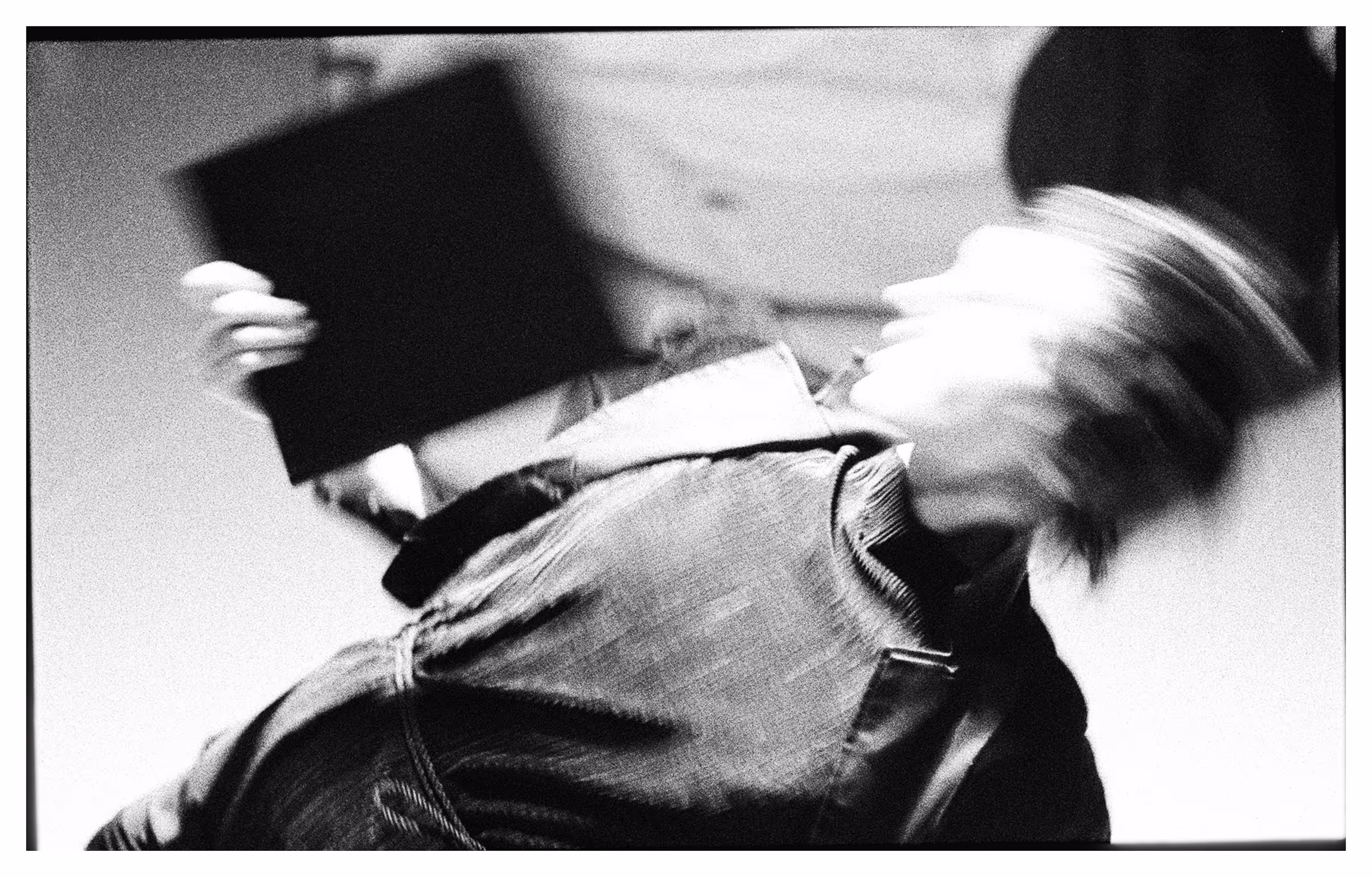 Black and white blurred photo of a person in a denim jacket swinging their head and holding a dark rectangular object.