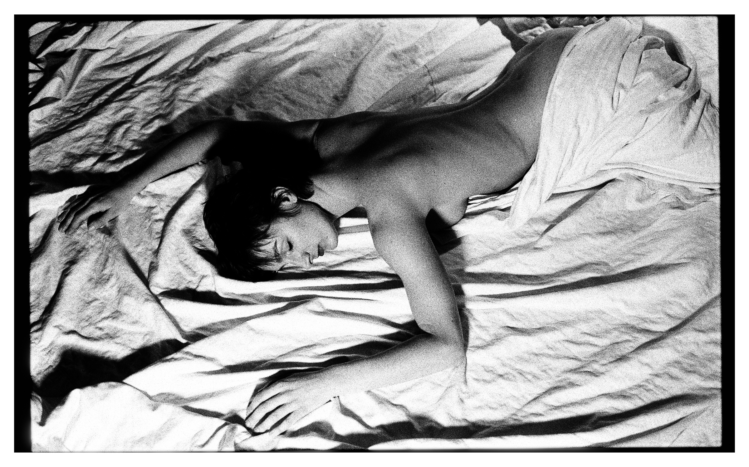 Black and white photo of a topless person lying face down on a wrinkled sheet, partially covered with fabric.