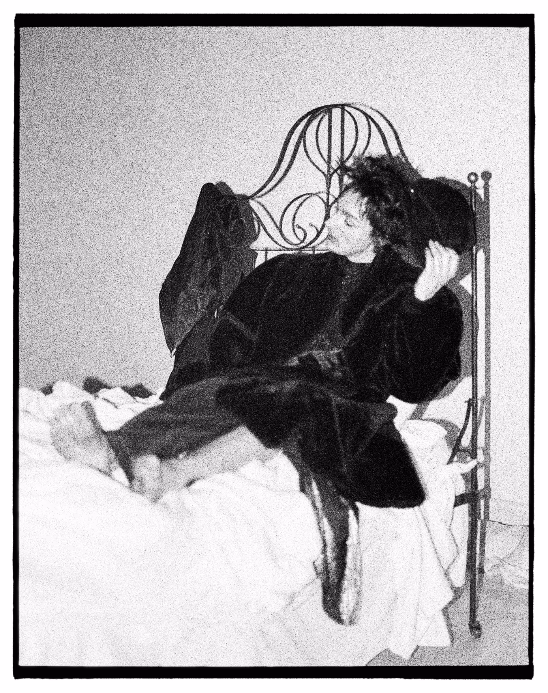 Black and white photo of a person reclining on a bed, wearing a dark fur coat and holding a hat, with a wrought iron headboard behind them.