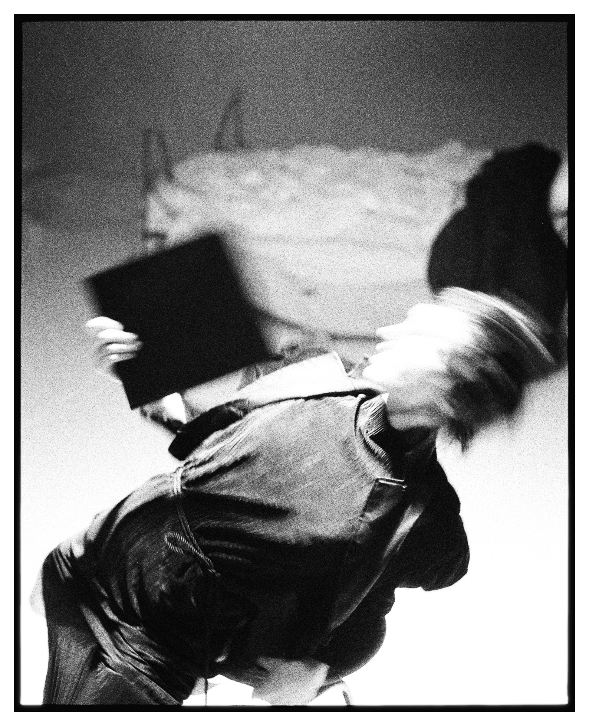 Black and white blurred image of a person in motion holding a dark rectangular object, with an unmade bed in the background.