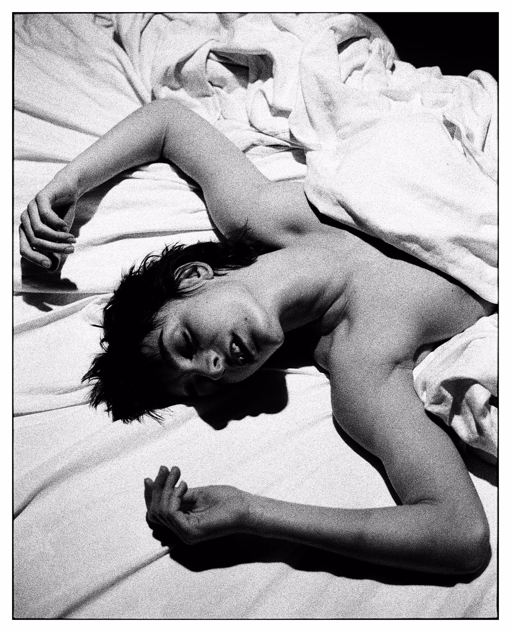 Black and white photo of a shirtless person lying on a bed with white sheets, eyes closed, and arms resting above and below their head.