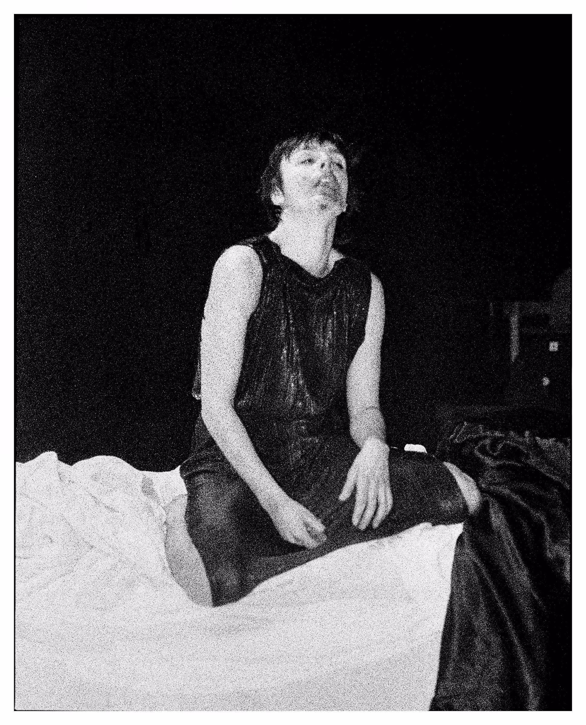 Black and white photo of a person sitting cross-legged on a bed with white sheets, wearing a sleeveless top.