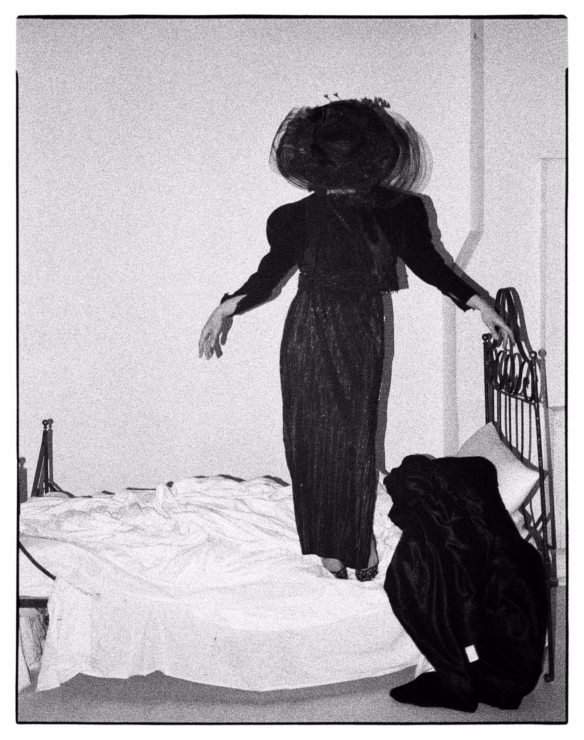 Black-and-white photo of a person in a long dark dress and large hat standing on an unmade bed facing away from the camera.