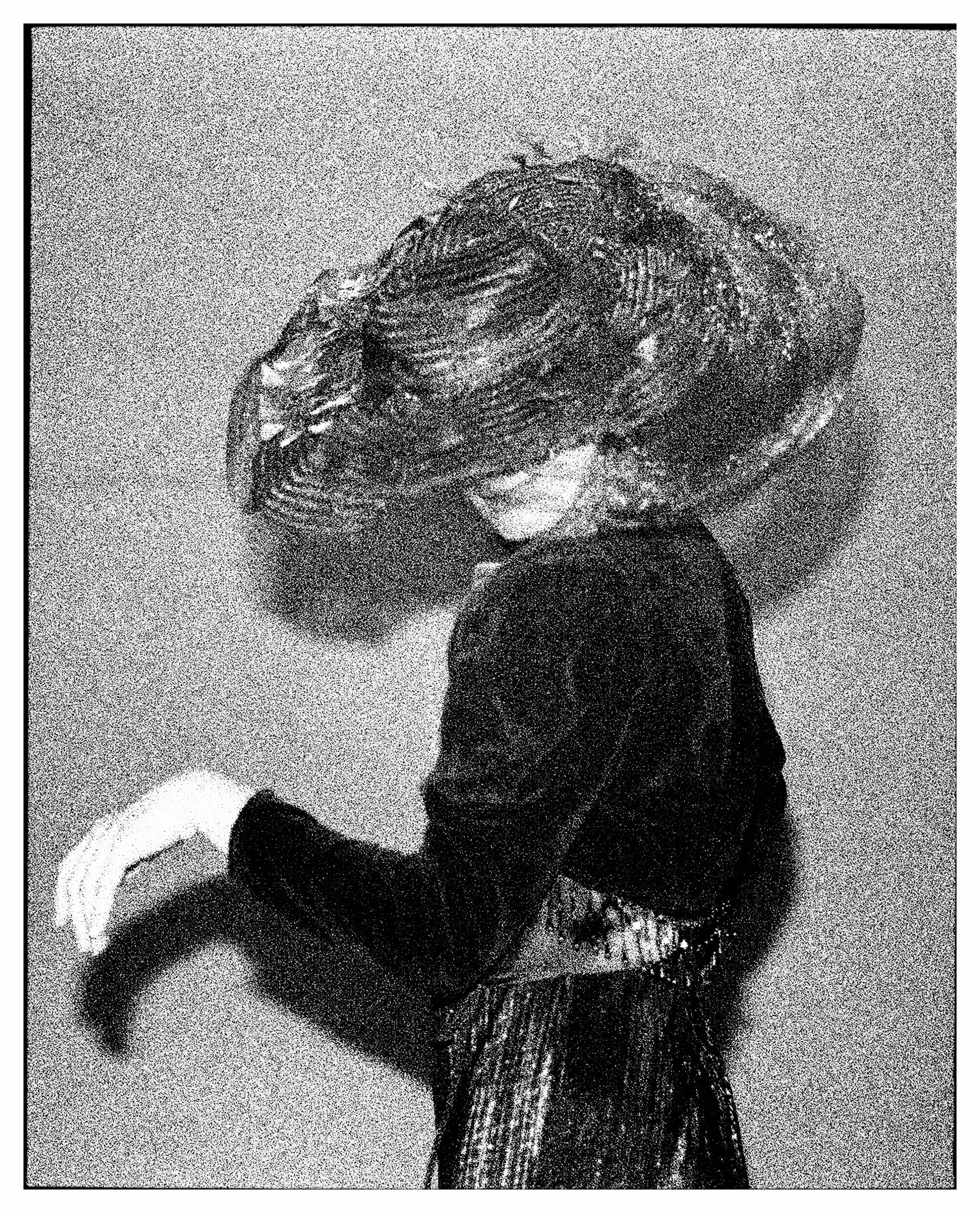 Black and white photo of a woman in vintage attire wearing a large ornate hat, posing sideways with one arm raised.