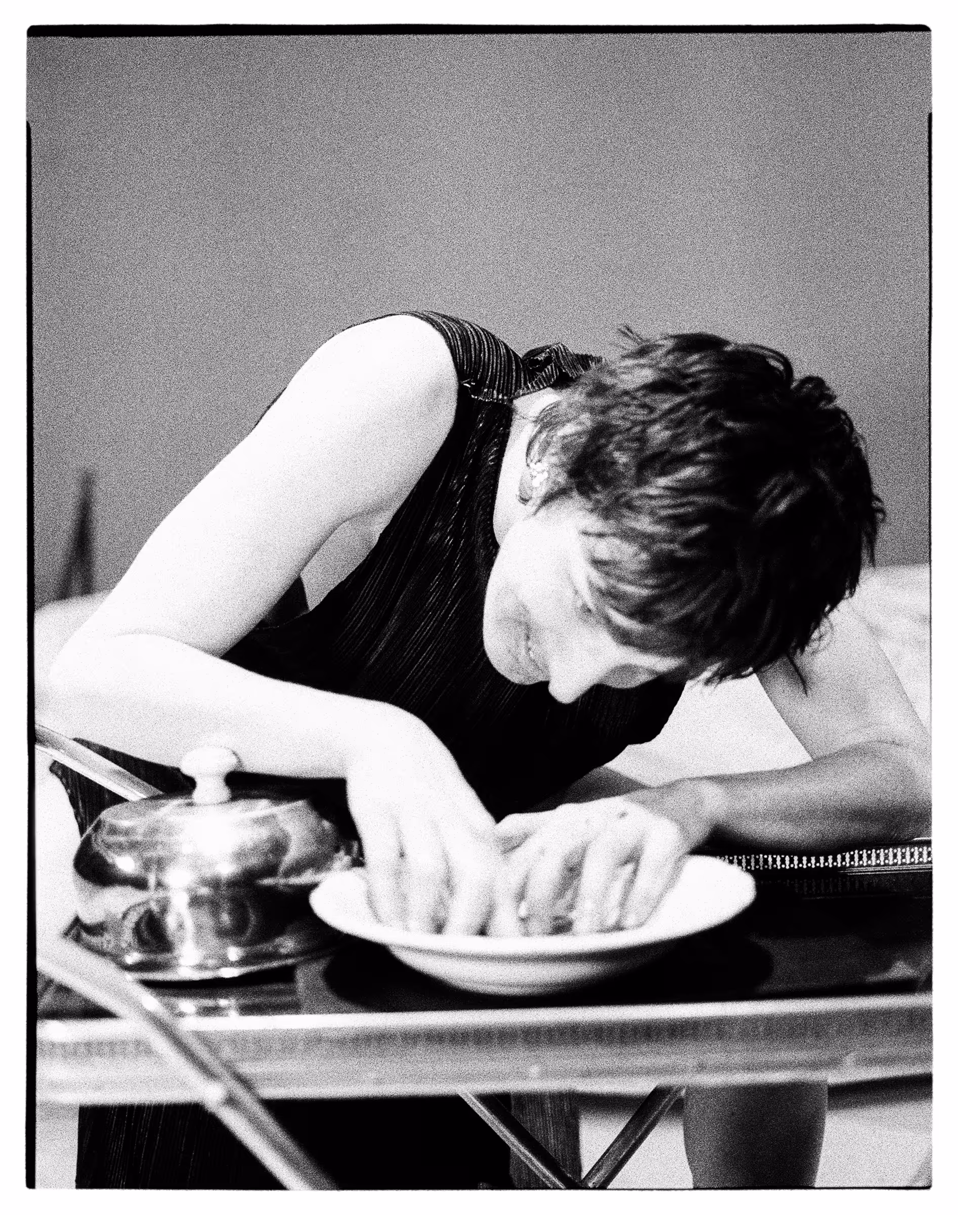 Black and white photo of a woman leaning over a table, smiling and reaching into a shallow bowl beside a covered dish.