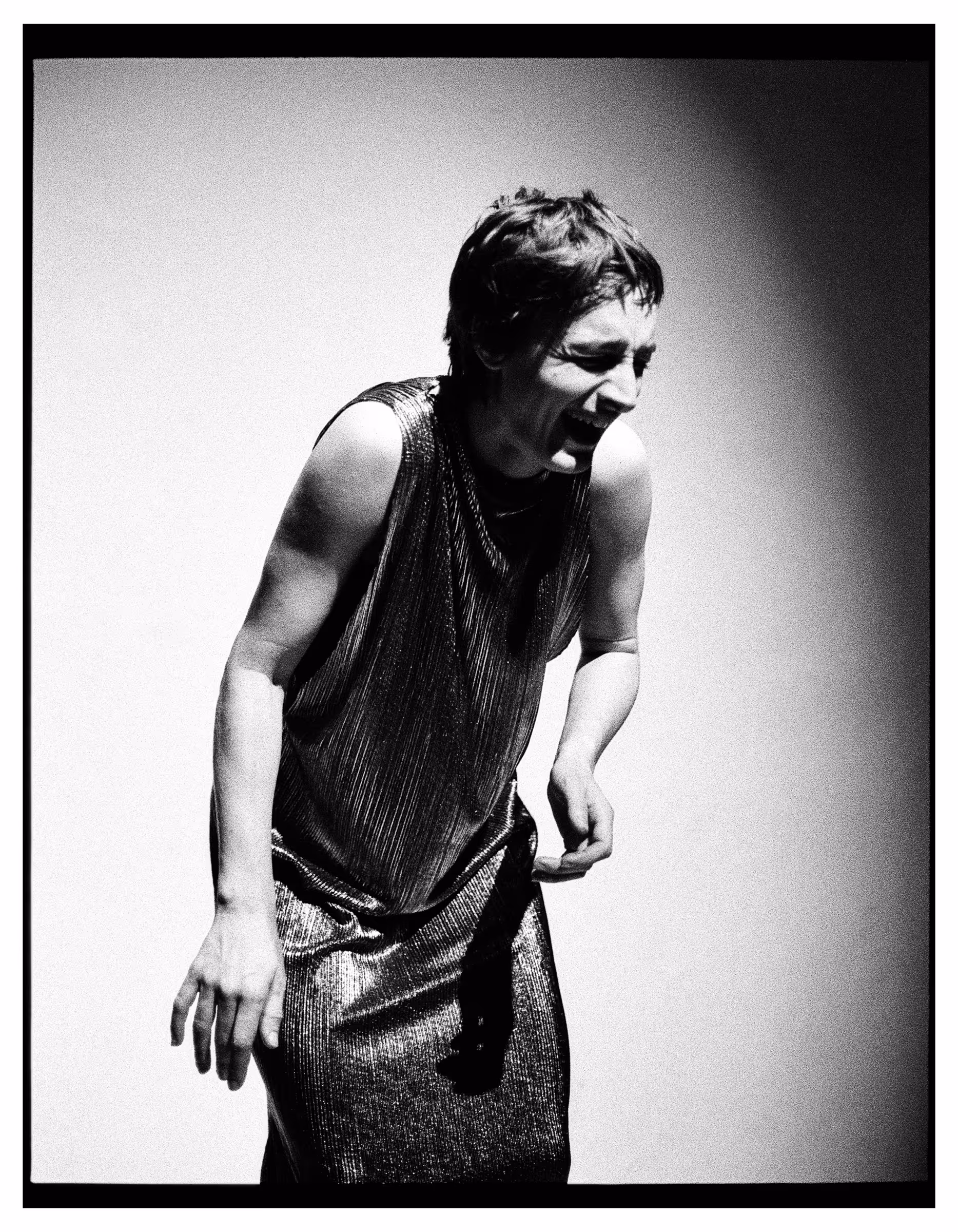 Black and white portrait of a person with short hair laughing and leaning forward wearing a sleeveless shiny textured dress.