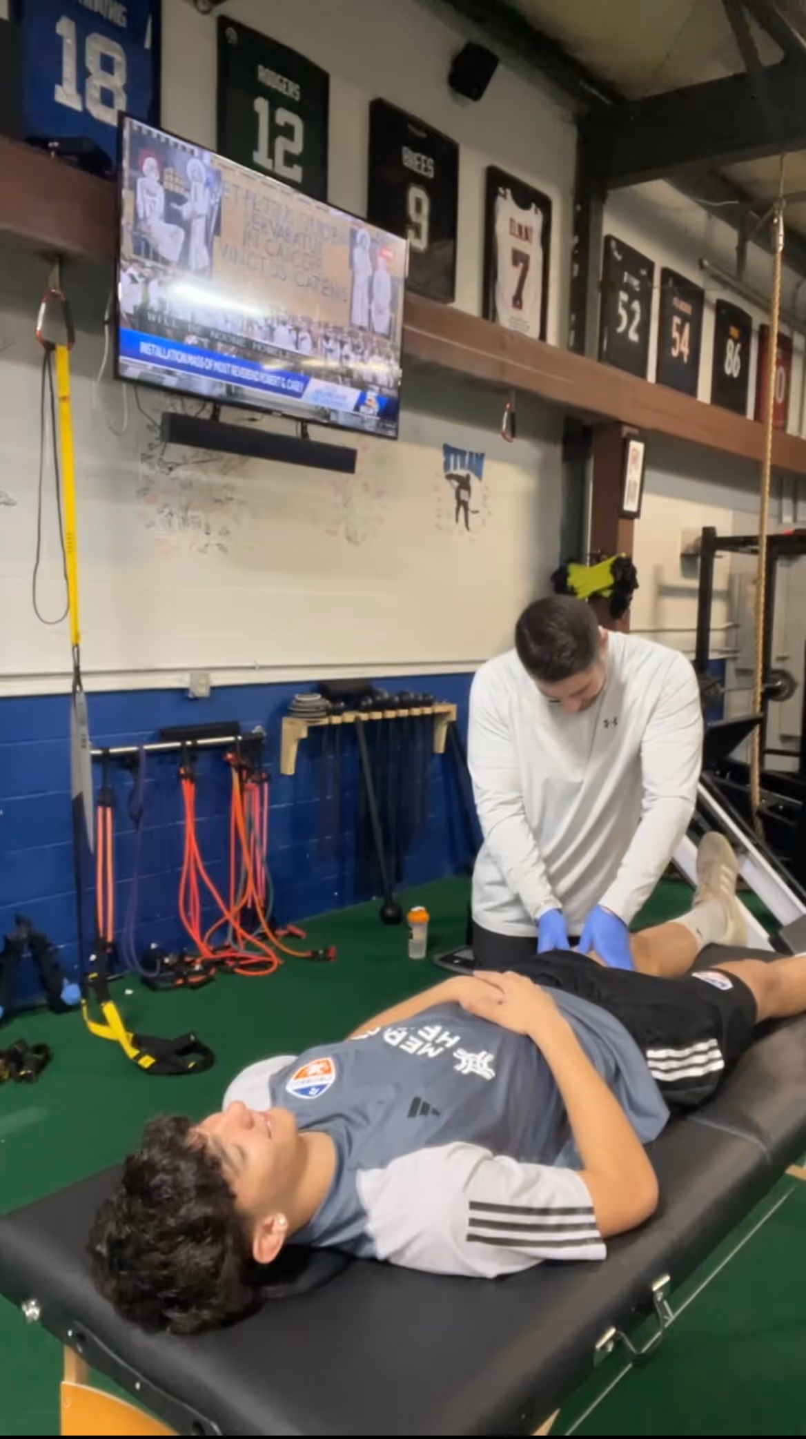 Athlete lying on a treatment table while a therapist wearing blue gloves examines his hip in a gym with sports jerseys and training equipment on the walls.