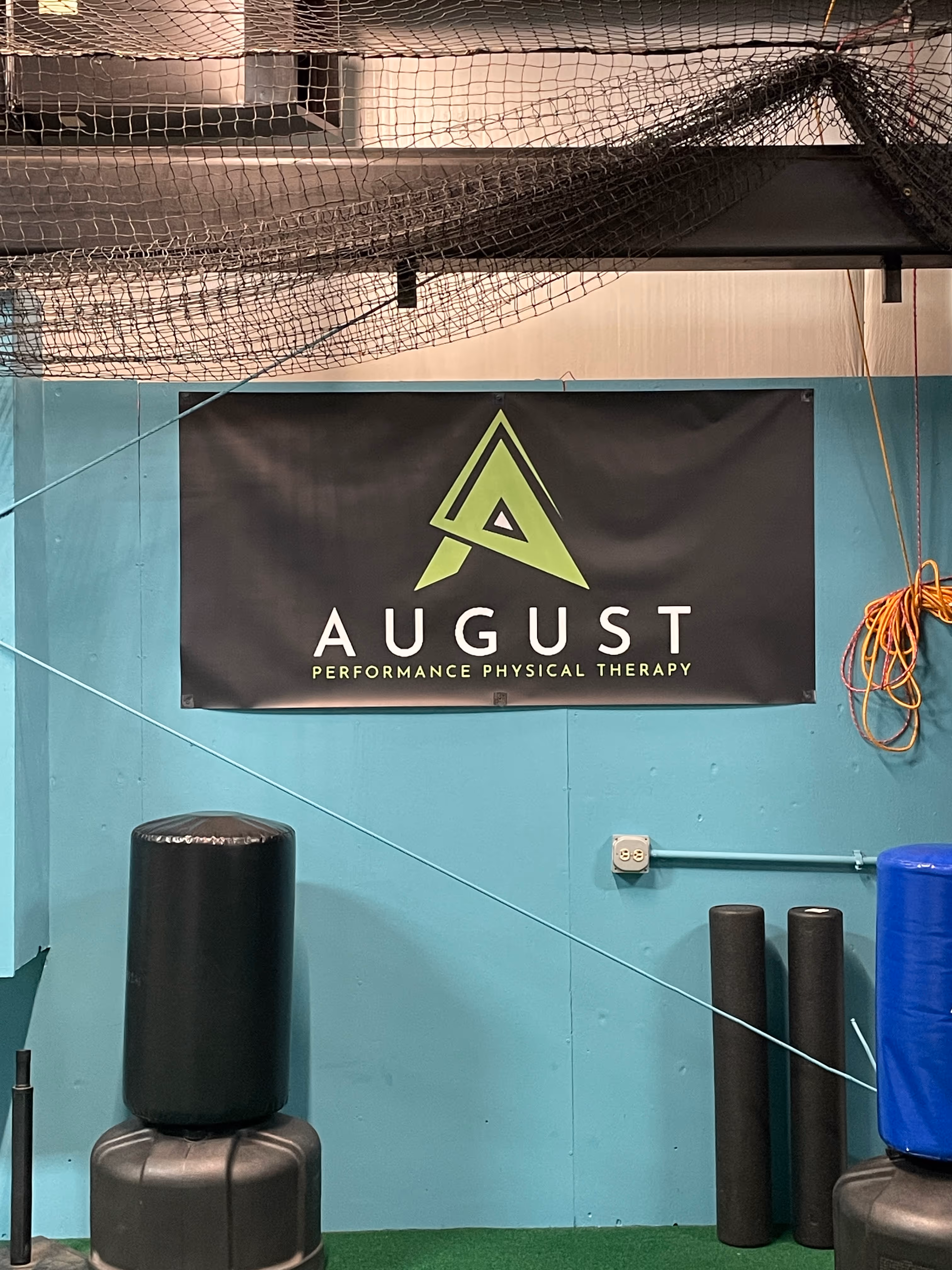 Indoor gym wall with a black banner displaying a green triangular logo and text 'AUGUST Performance Physical Therapy', surrounded by exercise equipment and a net overhead.