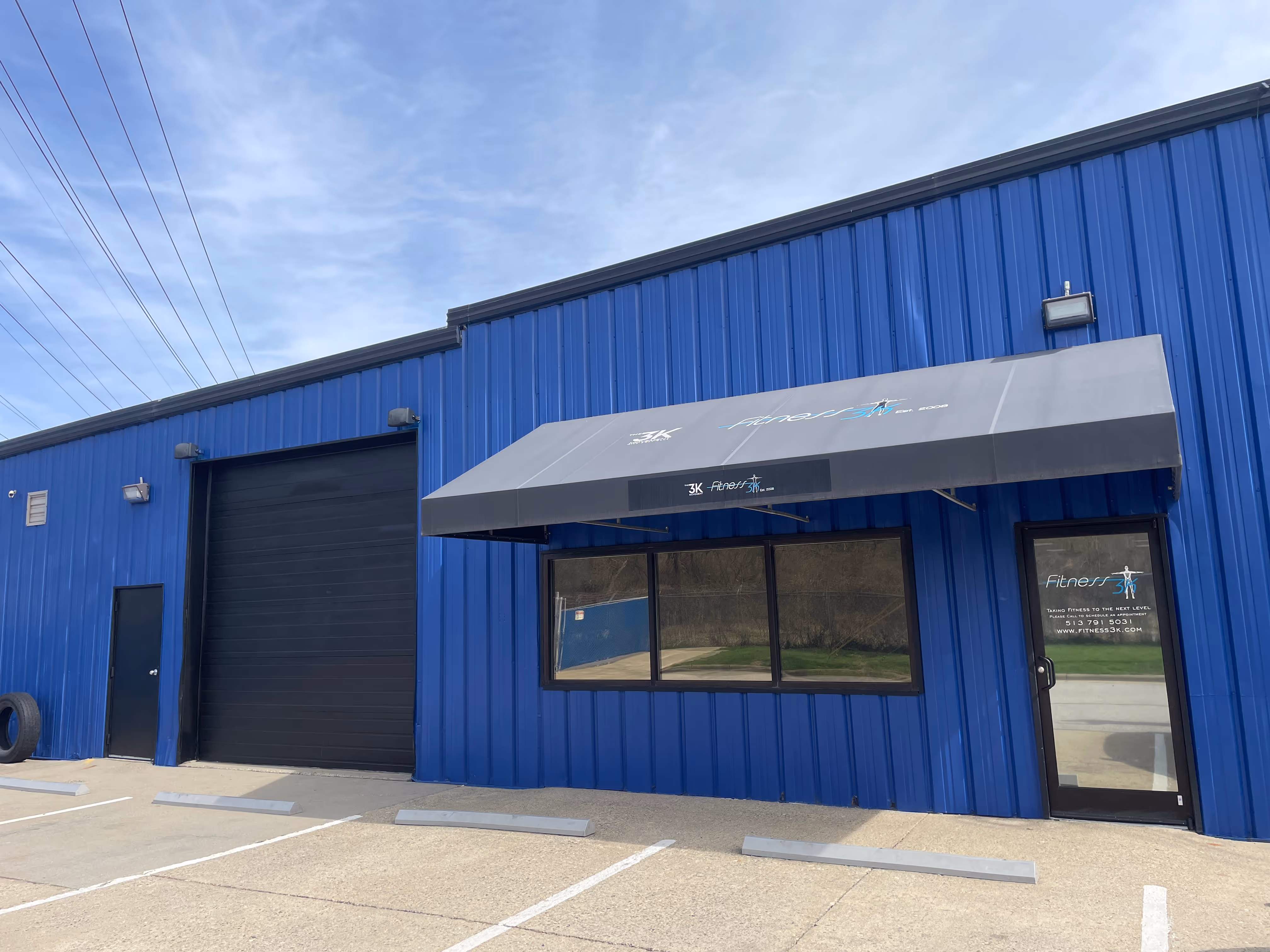 Blue building with a black garage door, black entrance door, and a black awning over a window and main entrance door with Fitness 3K signage.