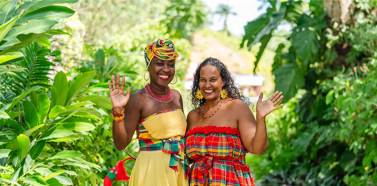 Two smiling women dressed in colorful traditional attire in a lush garden, raising their hands in greeting.