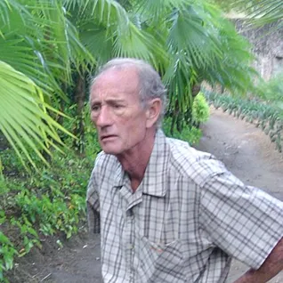 An elderly man wearing a plaid shirt, standing on a path surrounded by tropical vegetation.