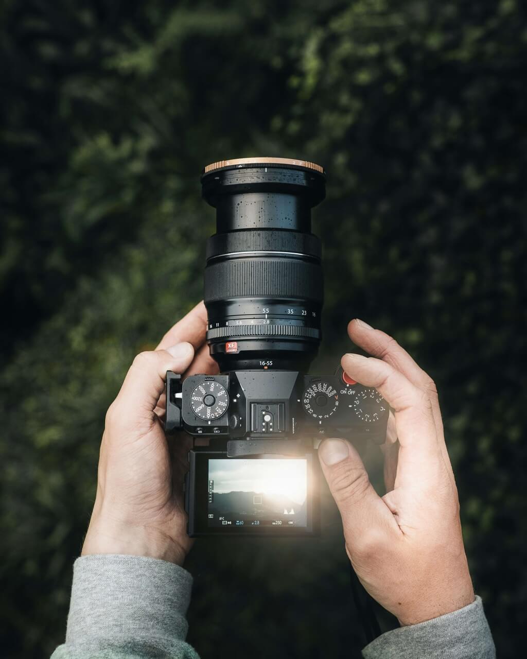 Close-up of hands holding a professional camera with a zoom lens, displaying a landscape with the sun on the LCD screen.
