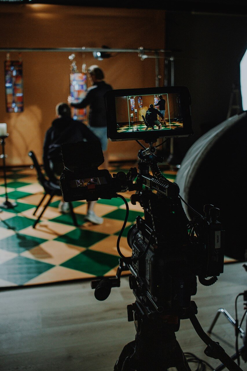 Camera filming two people in a studio with green and brown checkered floor and colorful hanging fabric panels.