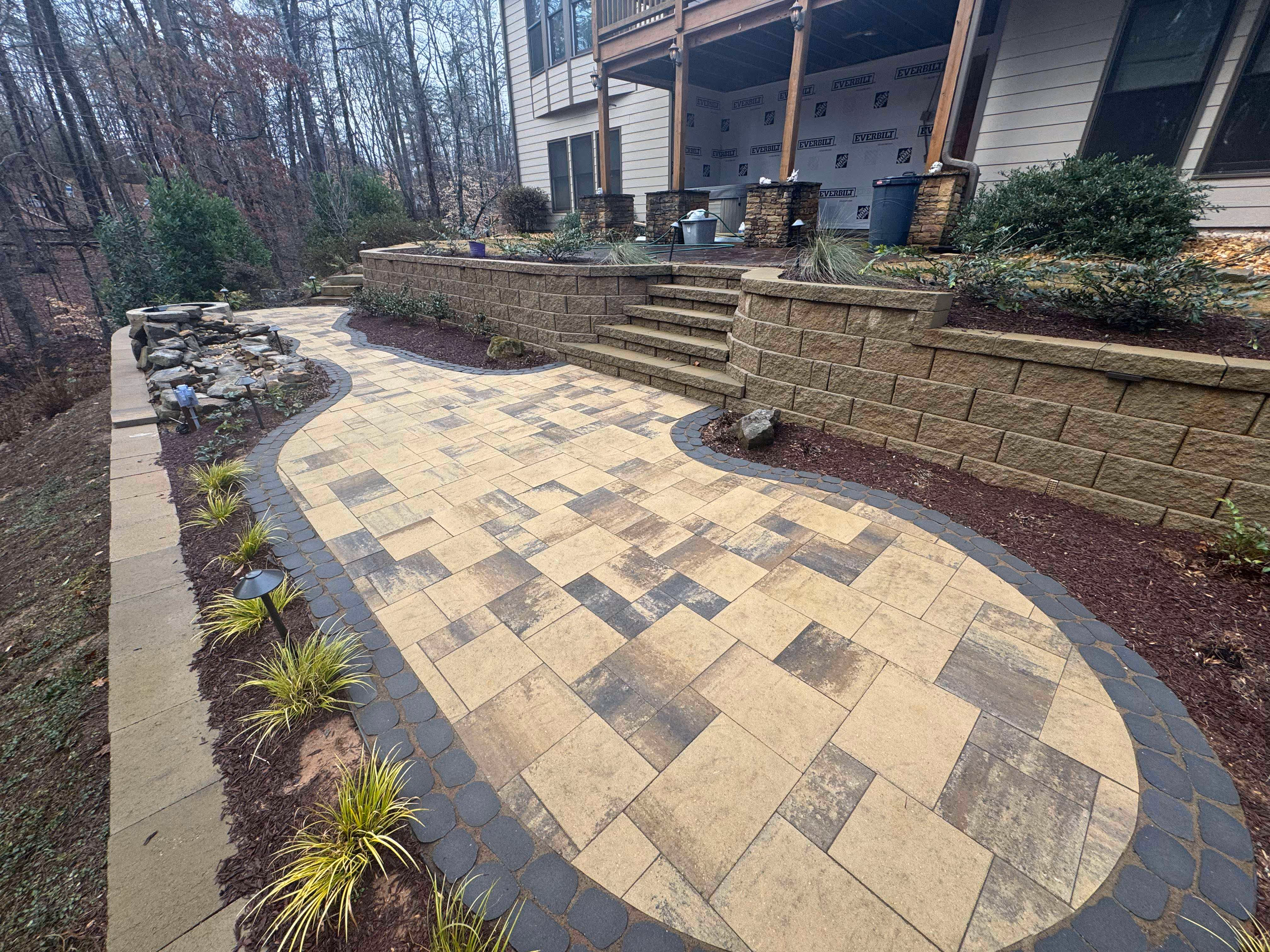 paver patio beneath modular retaining wall surrounded by mulch and plants with home in background
