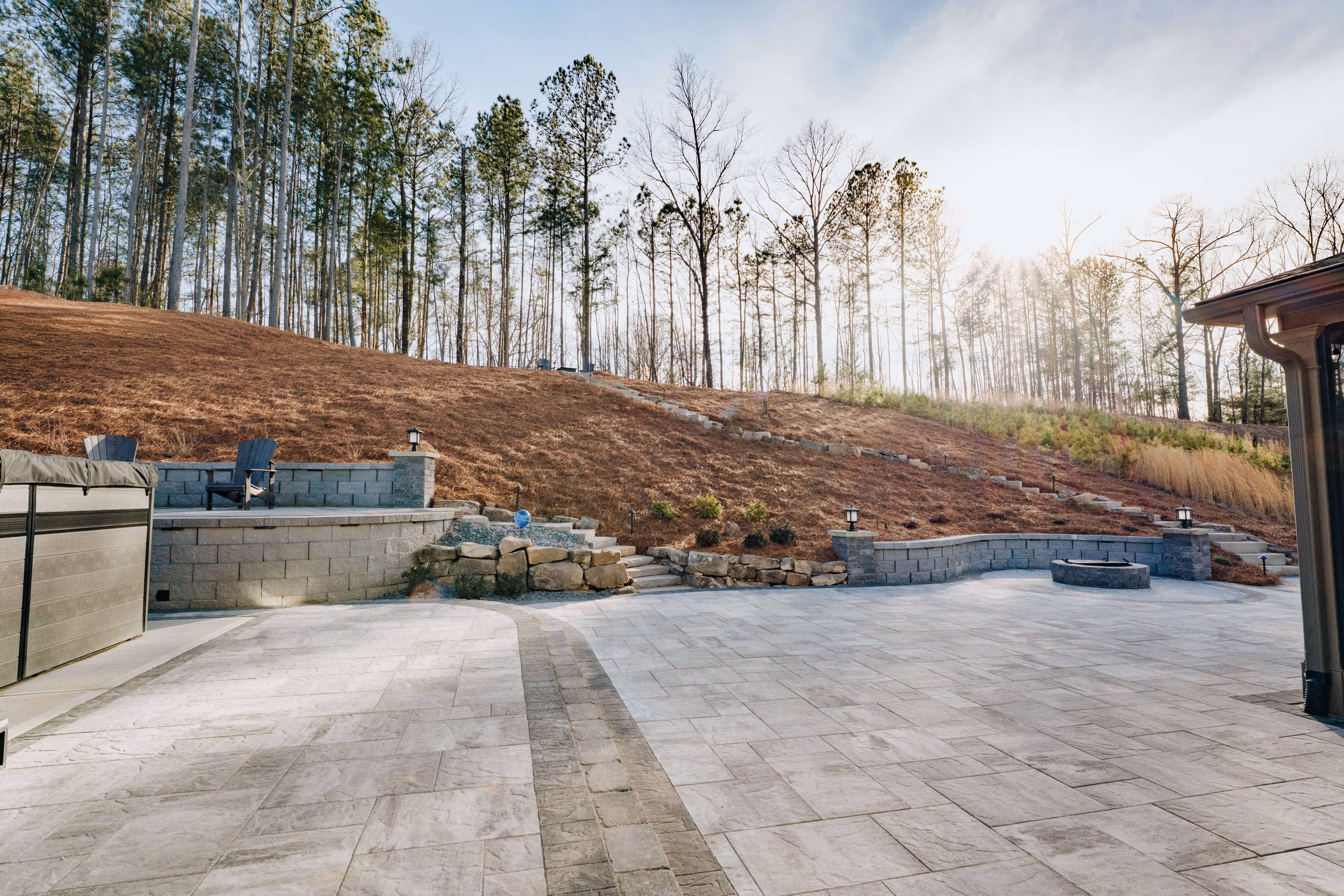 large paver patio with fire pit and seating area in front of pine straw slope and stone staircase