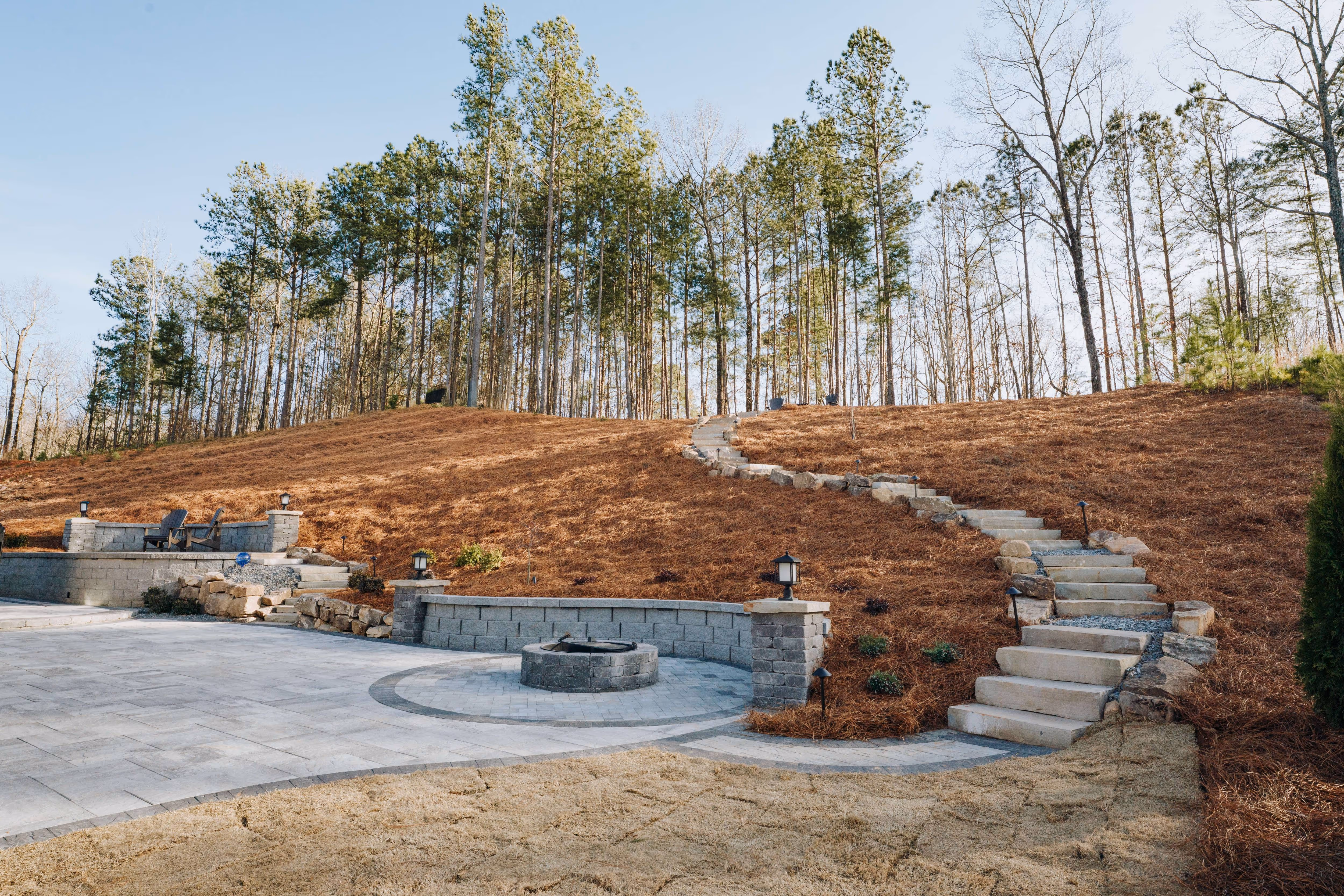 large paver patio with fire pit and seating area in front of pine straw slope and stone staircase