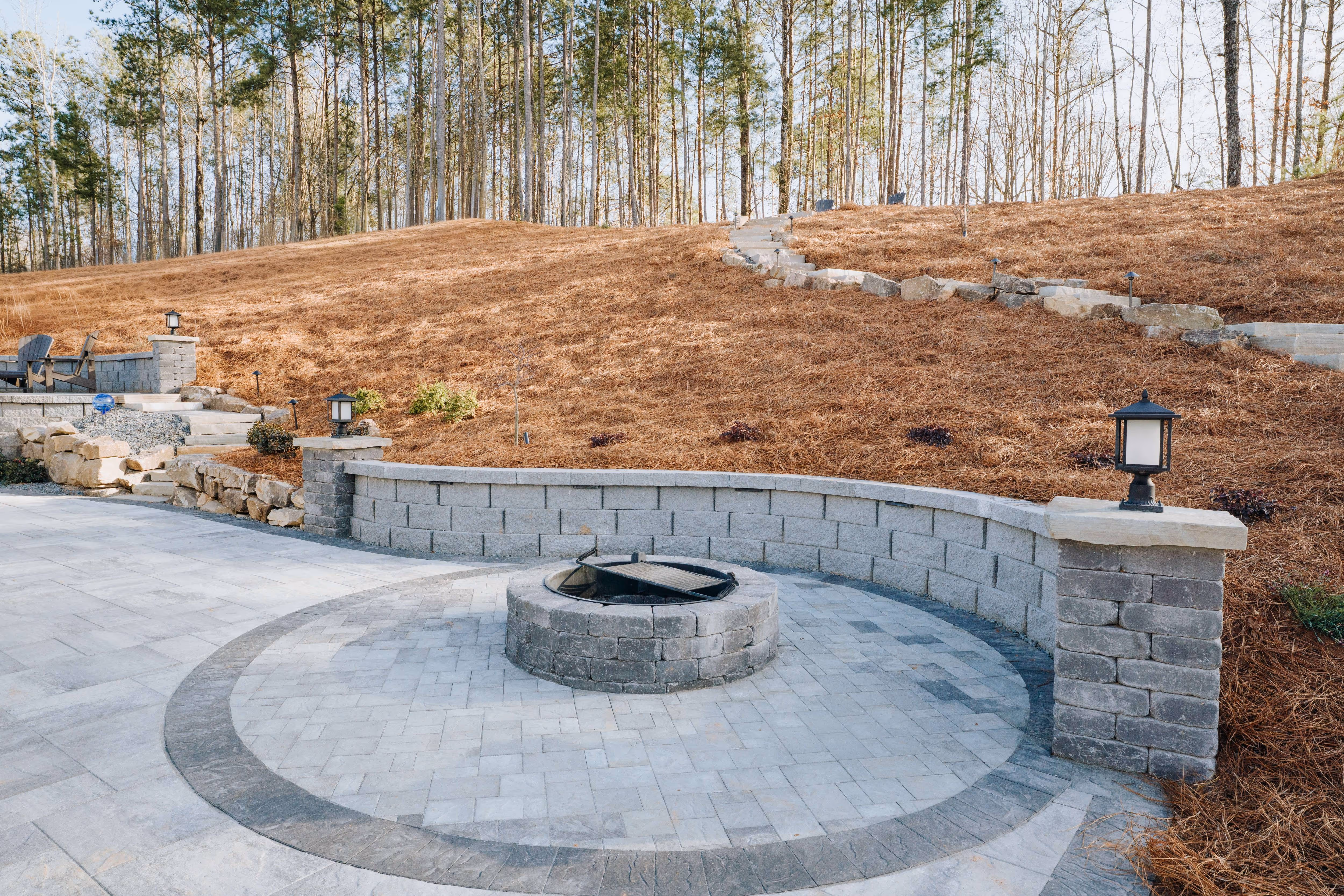 paver patio with retaining wall and fire pit with staircase and pine straw slope in background
