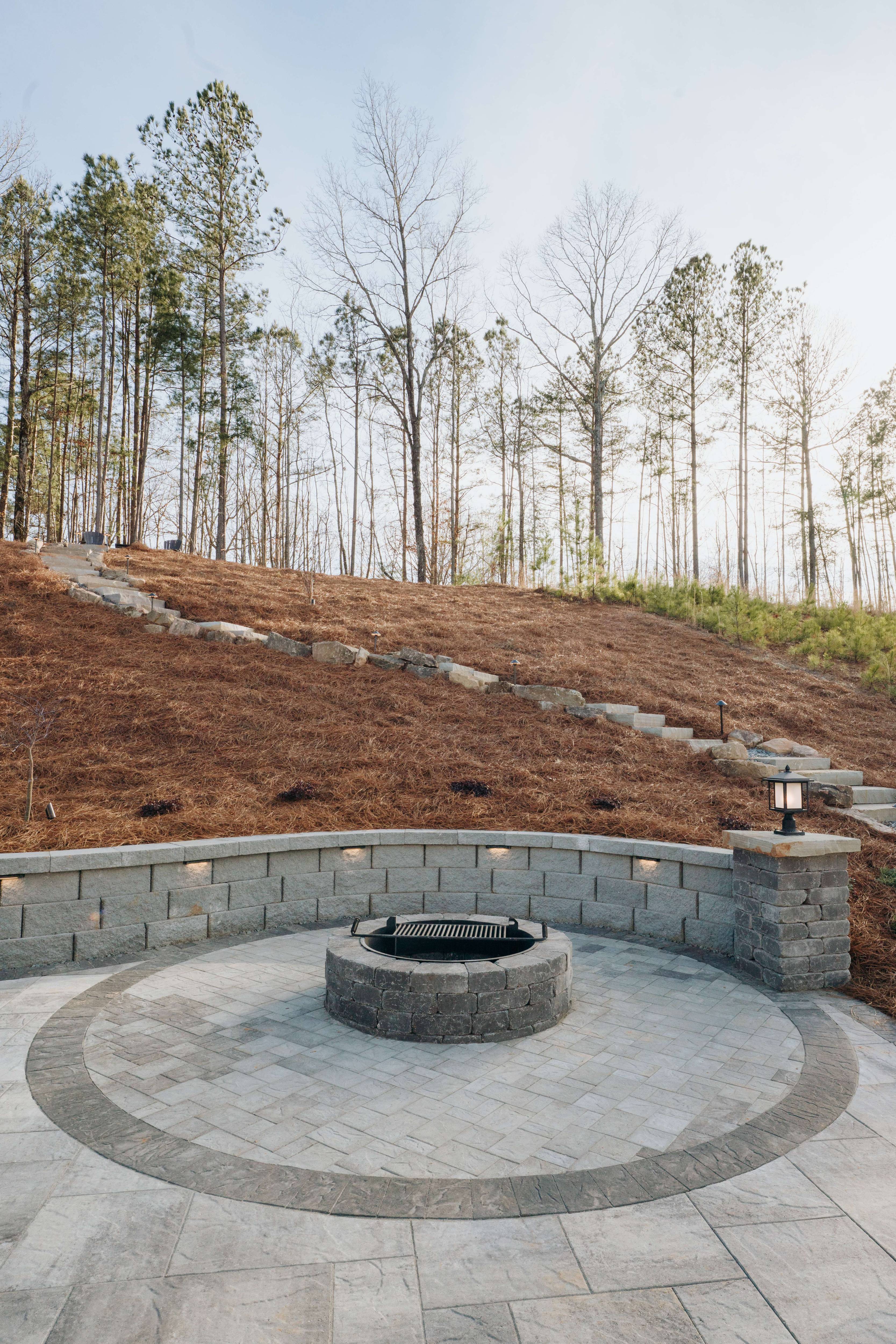 Stone patio with built-in fire pit and curved retaining wall with landscape lighting, surrounded by a pine forest in North Georgia