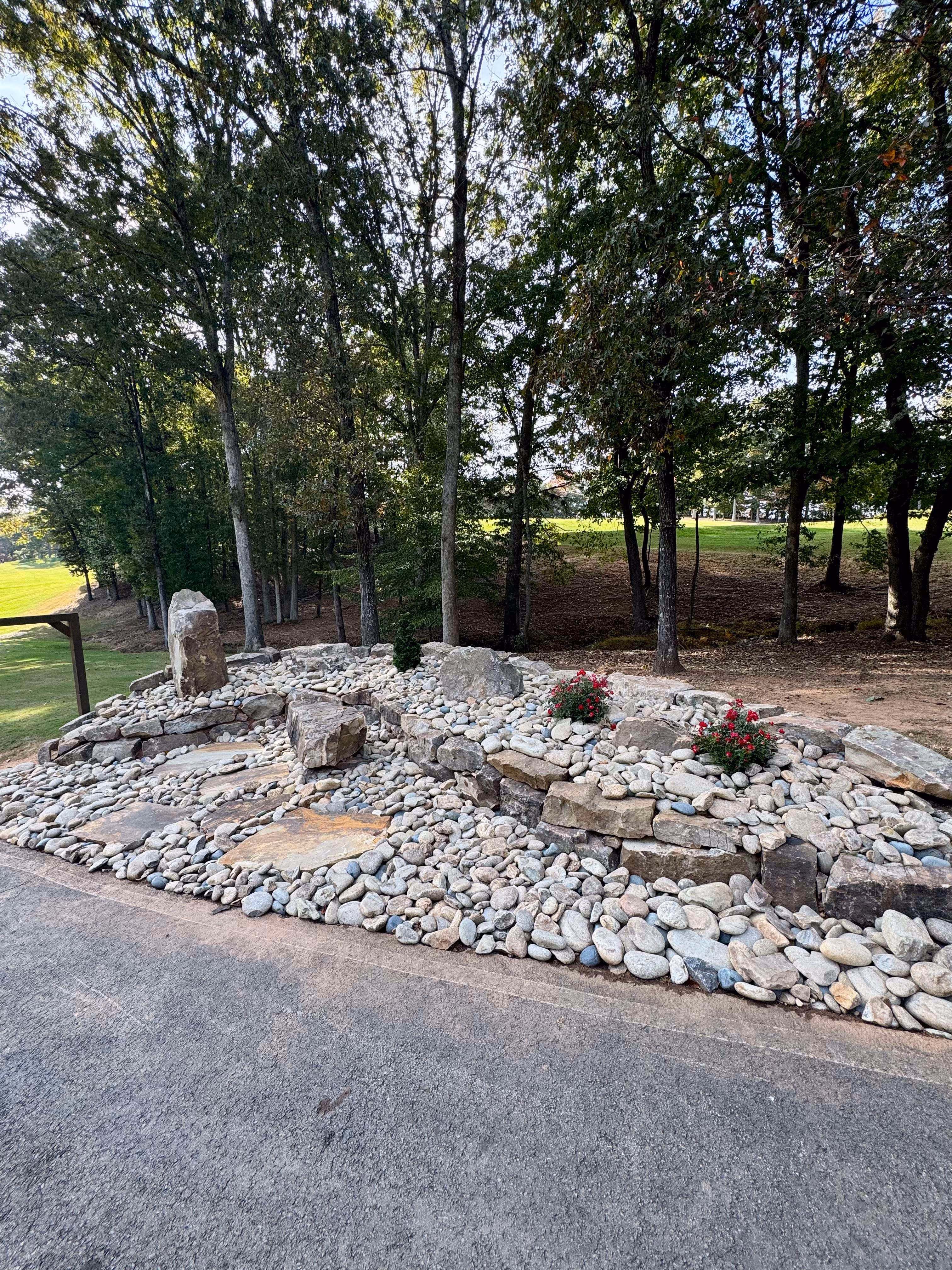River rock landscape bed with natural stone boulders and red flowering plants along a driveway edge