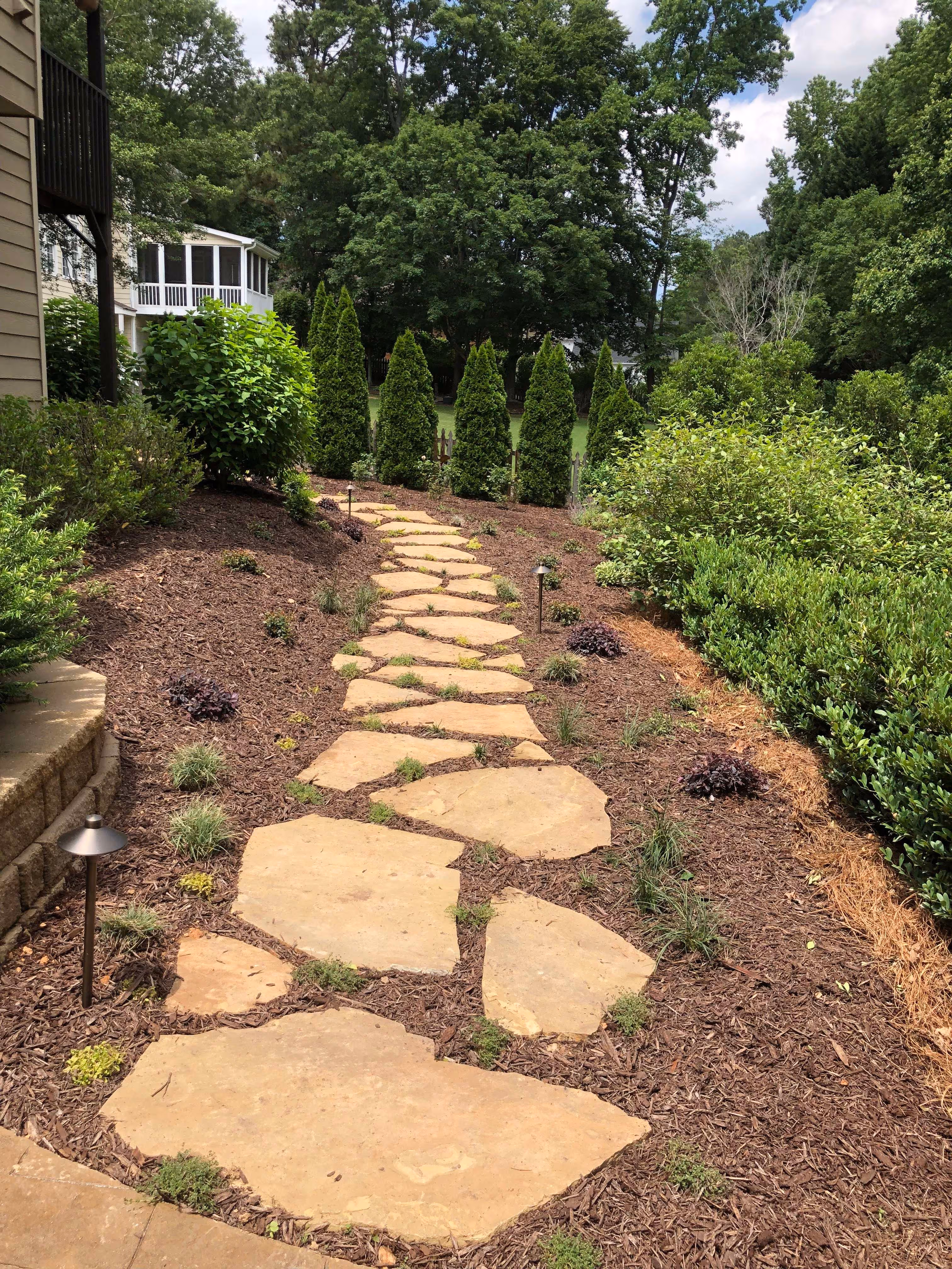 flagstone walkway in middle of pine straw area with trees and bushes in background