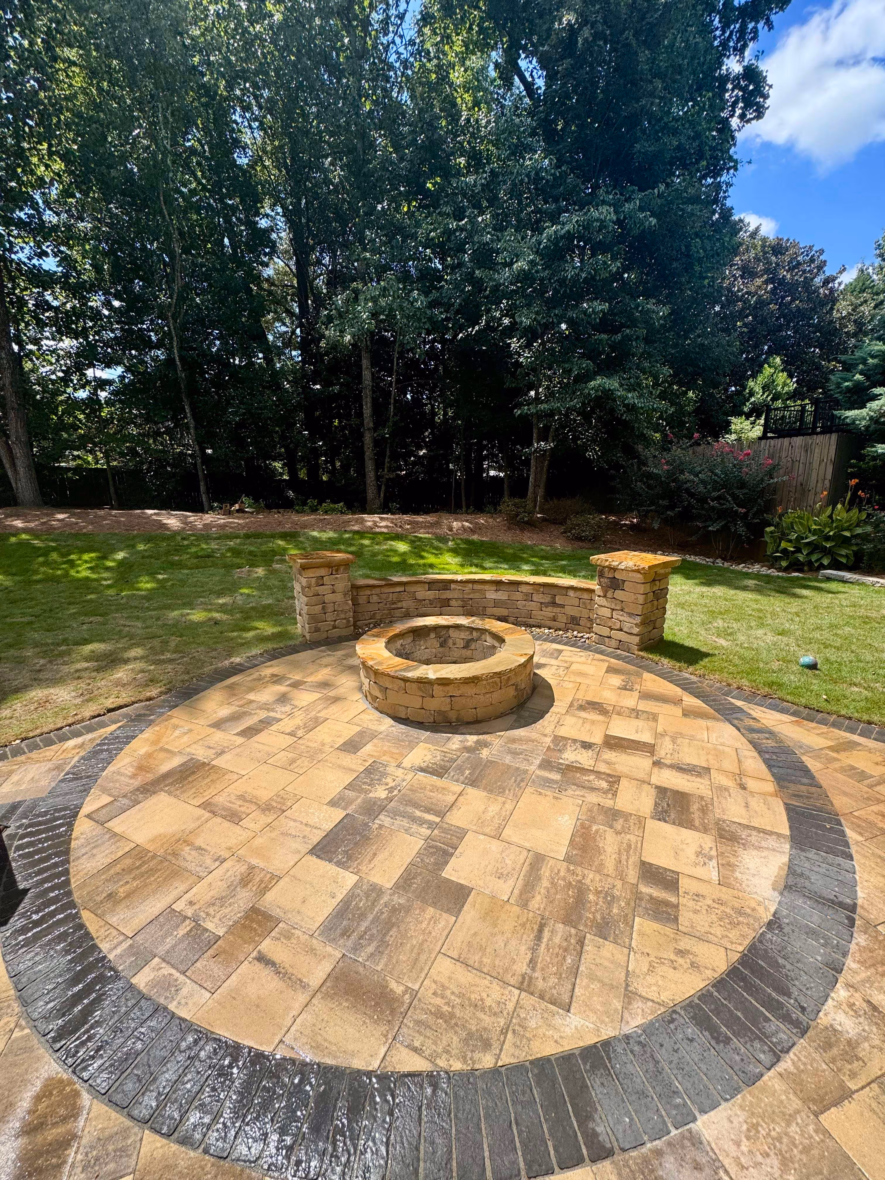 paver patio with fire pit in the middle and grass and trees in background
