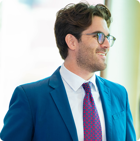 Smiling man with glasses wearing a blue suit, white shirt, and patterned red and blue tie.