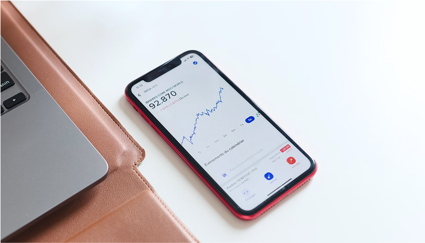 Smartphone displaying a financial stock graph beside a laptop on a beige leather surface.