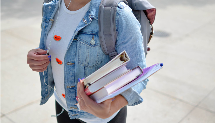 Student wearing denim jacket and backpack holding textbooks and notebooks while walking outdoors.