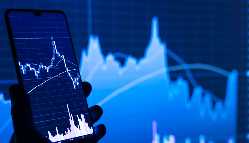 Hand holding smartphone showing financial candlestick chart against a large blue stock market graph background.