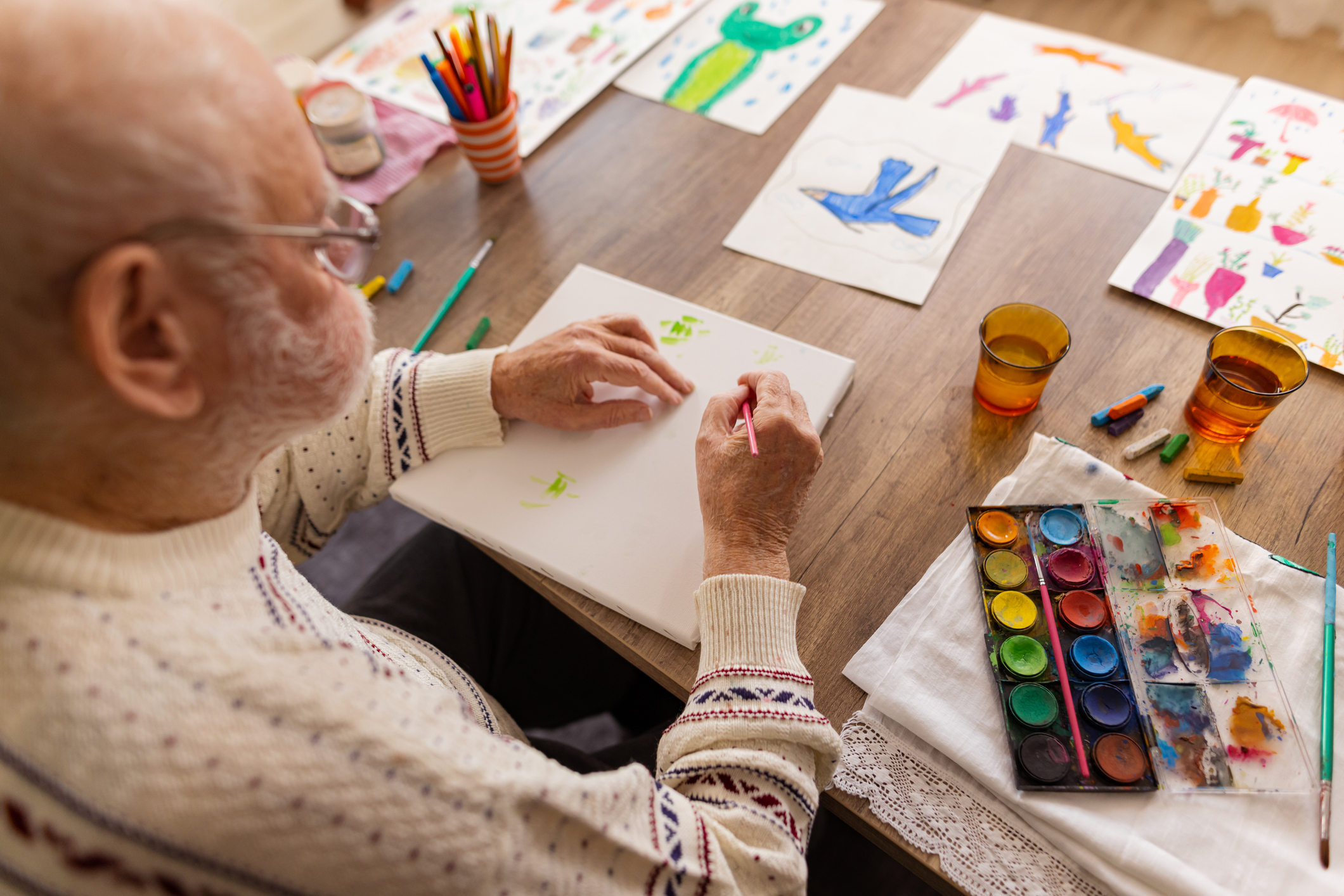 Elderly man painting with watercolors at a table filled with drawings and art supplies.