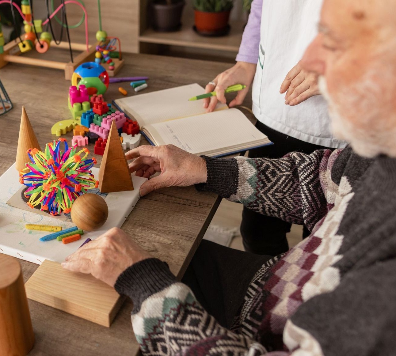 Elderly man working with wooden shapes and colorful toys on a table, assisted by a caregiver with a notebook.