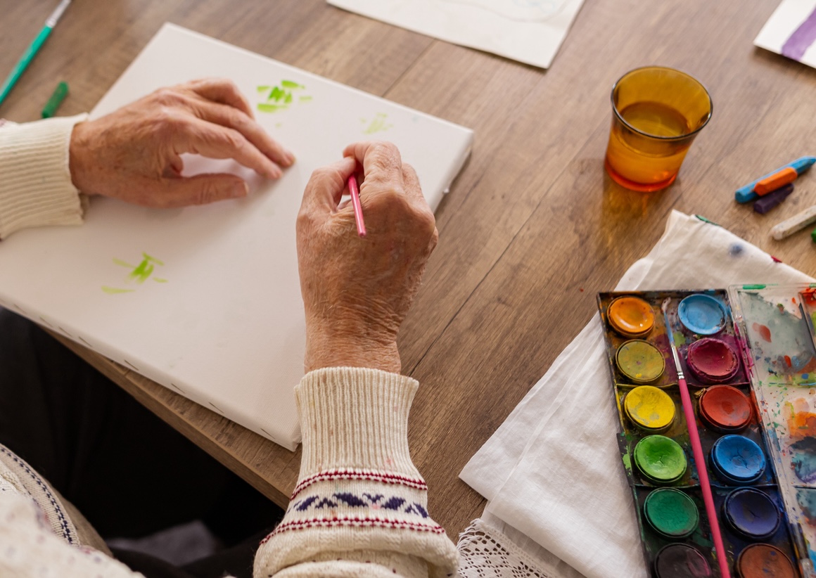 An elderly person painting on a canvas with watercolors on a wooden table.